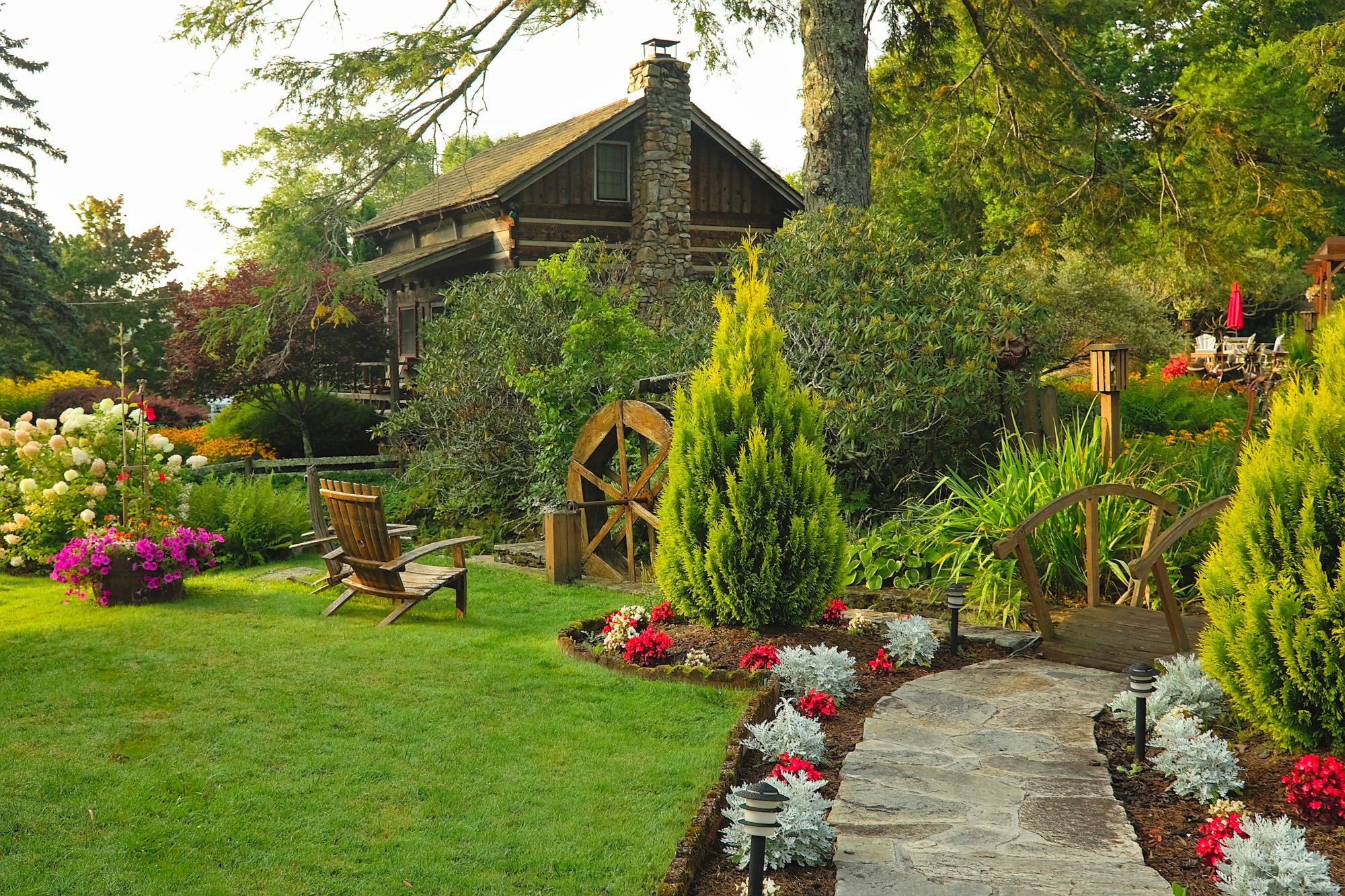 View of the log cabin from a distance, with a water wheel, flowers, and chairs in the foreground