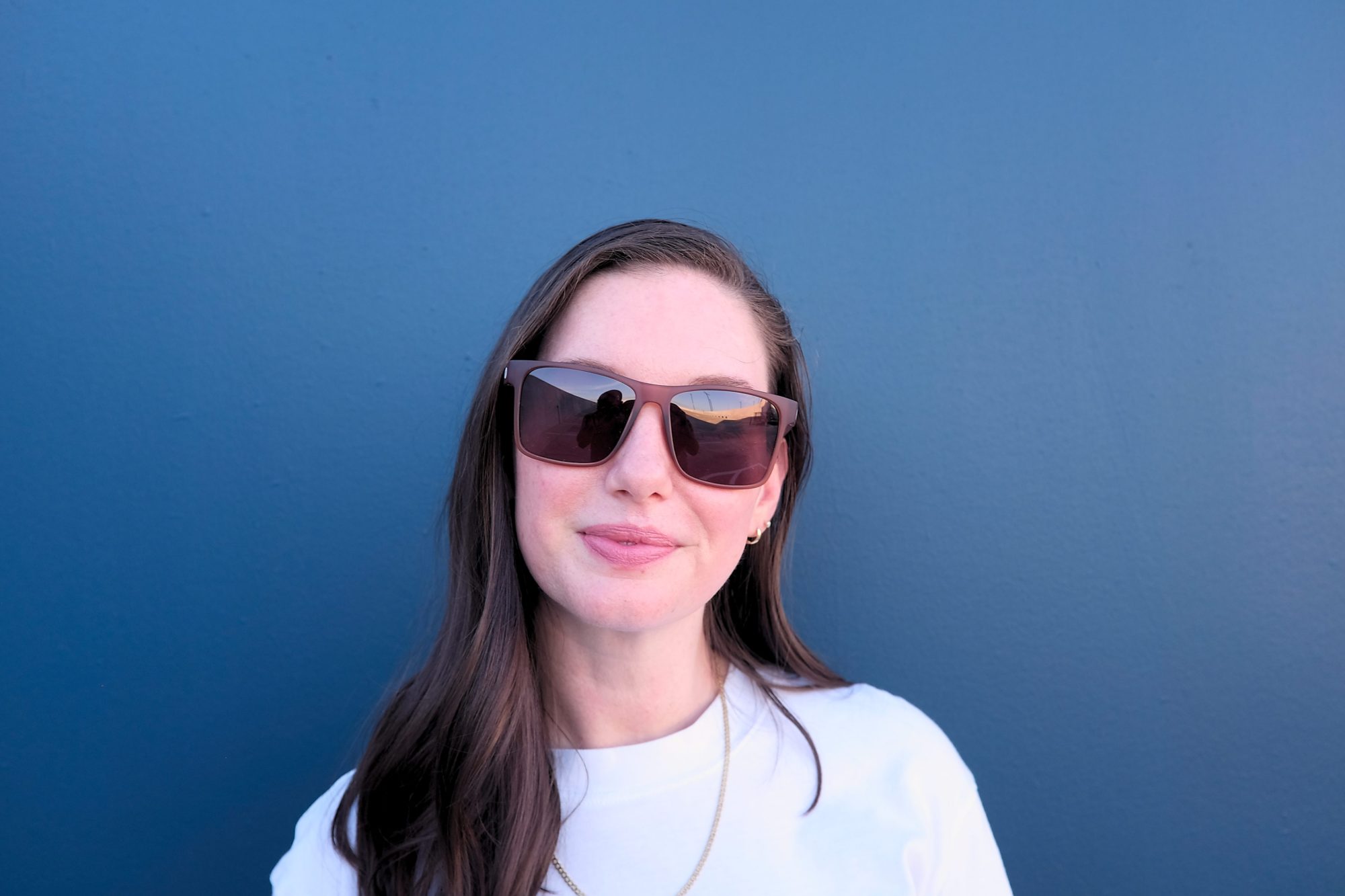 Alyssa wears the Sunski Puerto sunglasses in Sienna Ruby in front of a blue wall and Michael is reflected back in the glasses