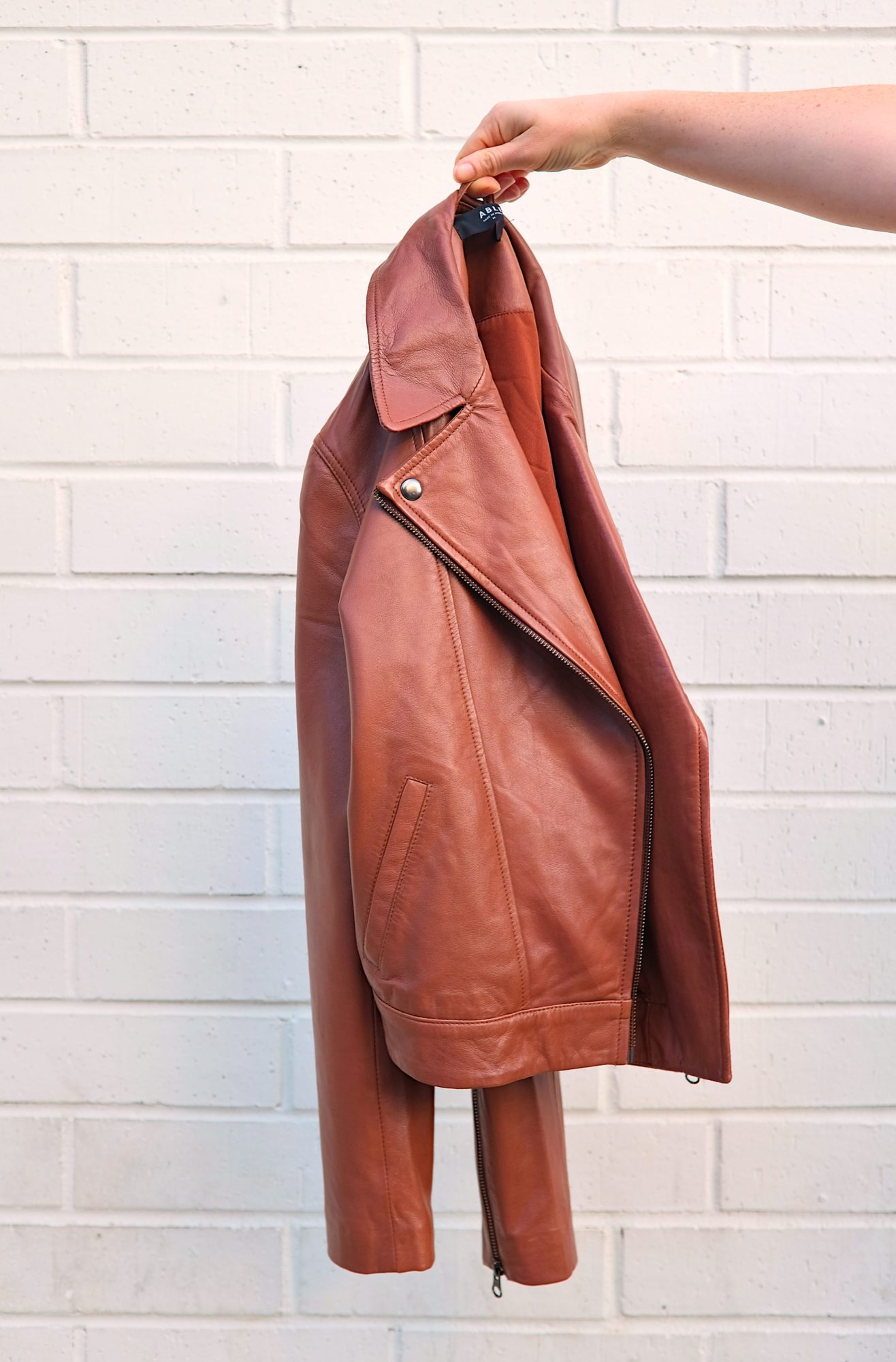 A hand holds a brown leather jacket from ABLE in front of a brick wall