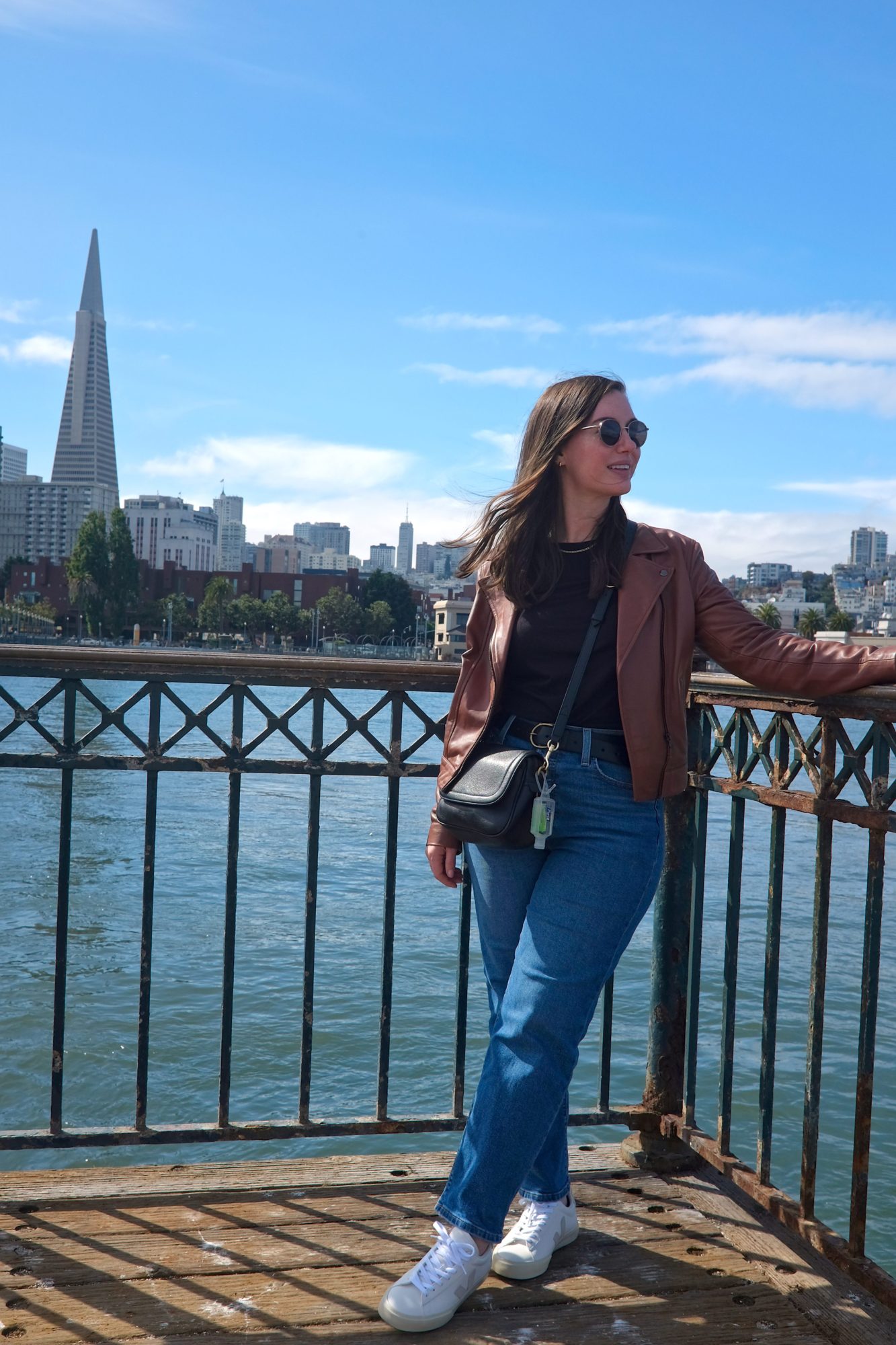Alyssa looks away from the camera while wearing a black top, brown jacket, blue jeans, and sneakers. San Francisco is in the background