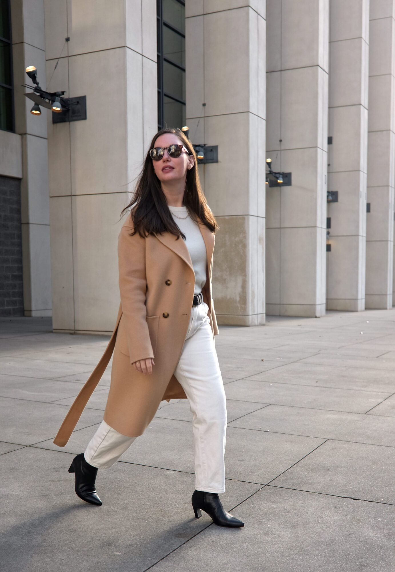 Alyssa wears a white top and jeans with black boots, a black belt, and camel coat