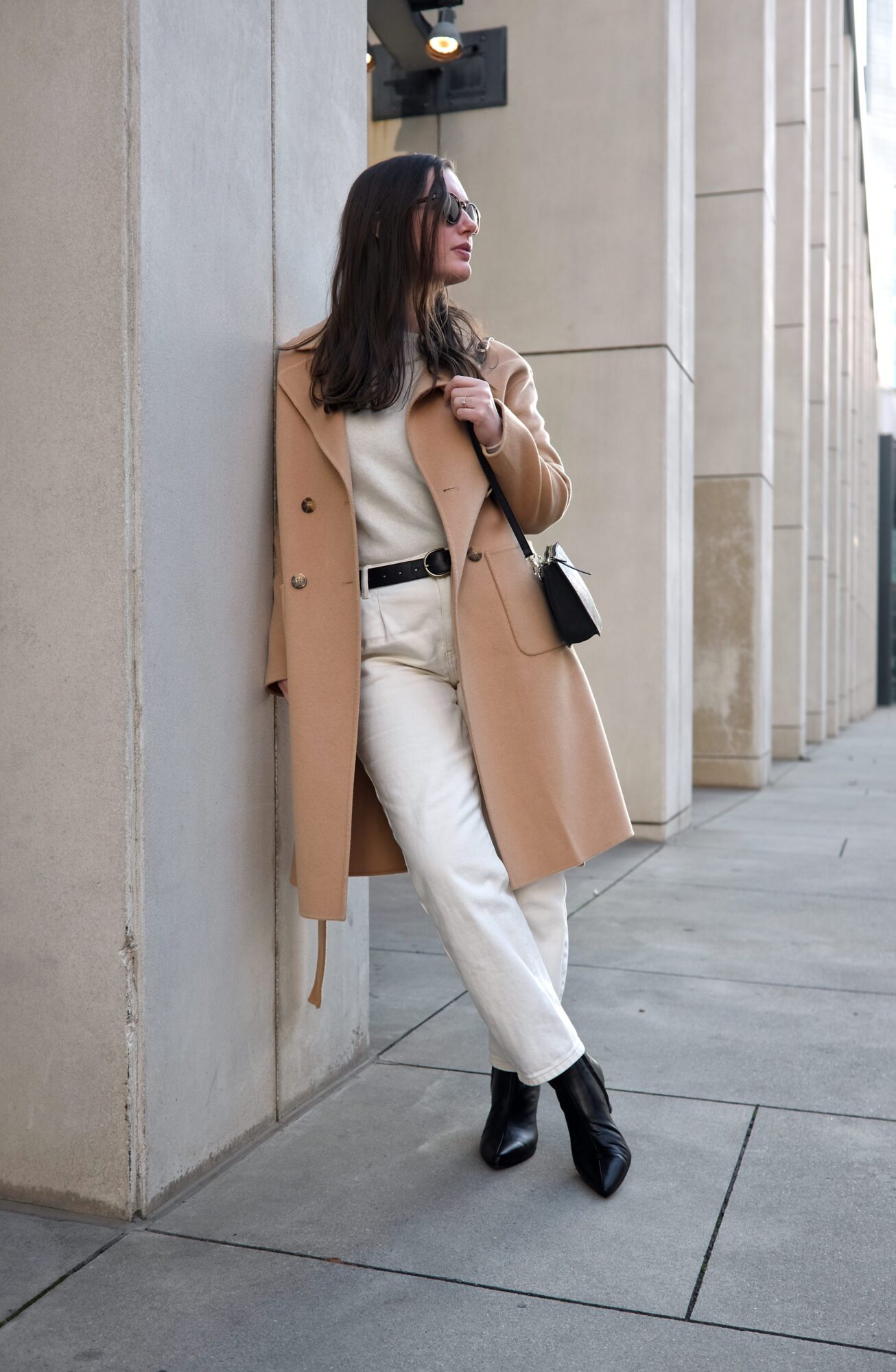 Alyssa wears a white top, white jeans, black boots, and camel coat and leans on a column