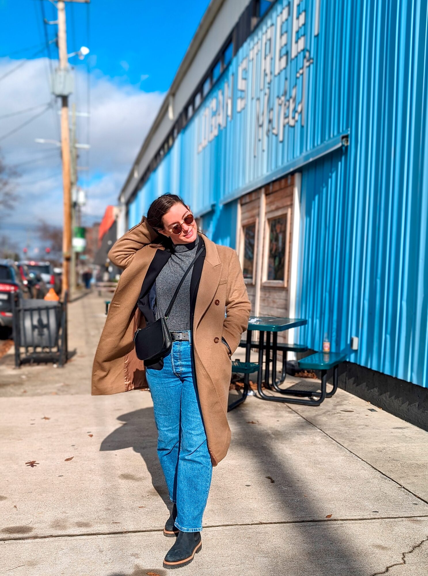 Alyssa wears a grey sweater with a camel coat and blue jeans in front of Louisville's Logan Street Market