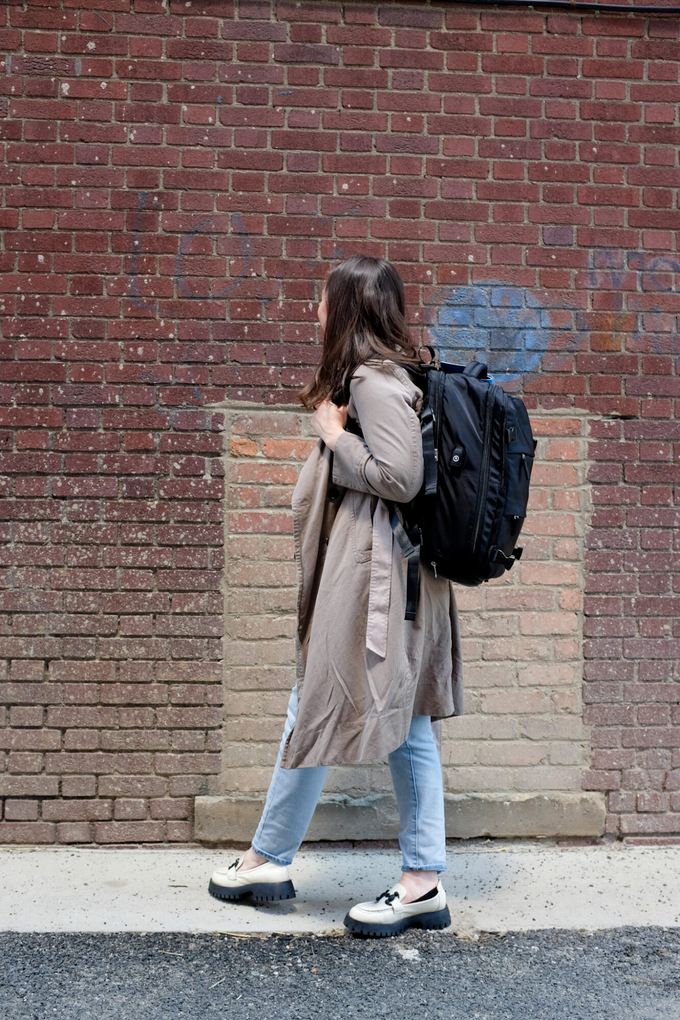 Alyssa wears a tan trench and carries a black backpack that fits Spirit Airlines' personal item dimensions