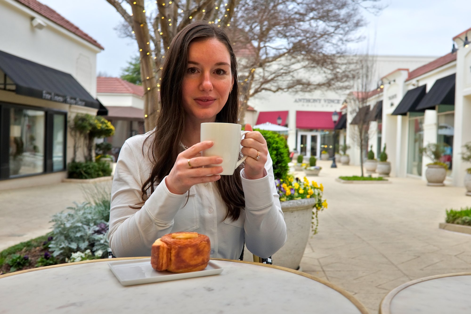 Alyssa sits at a bistro table with a pastry and holds a cup of coffee