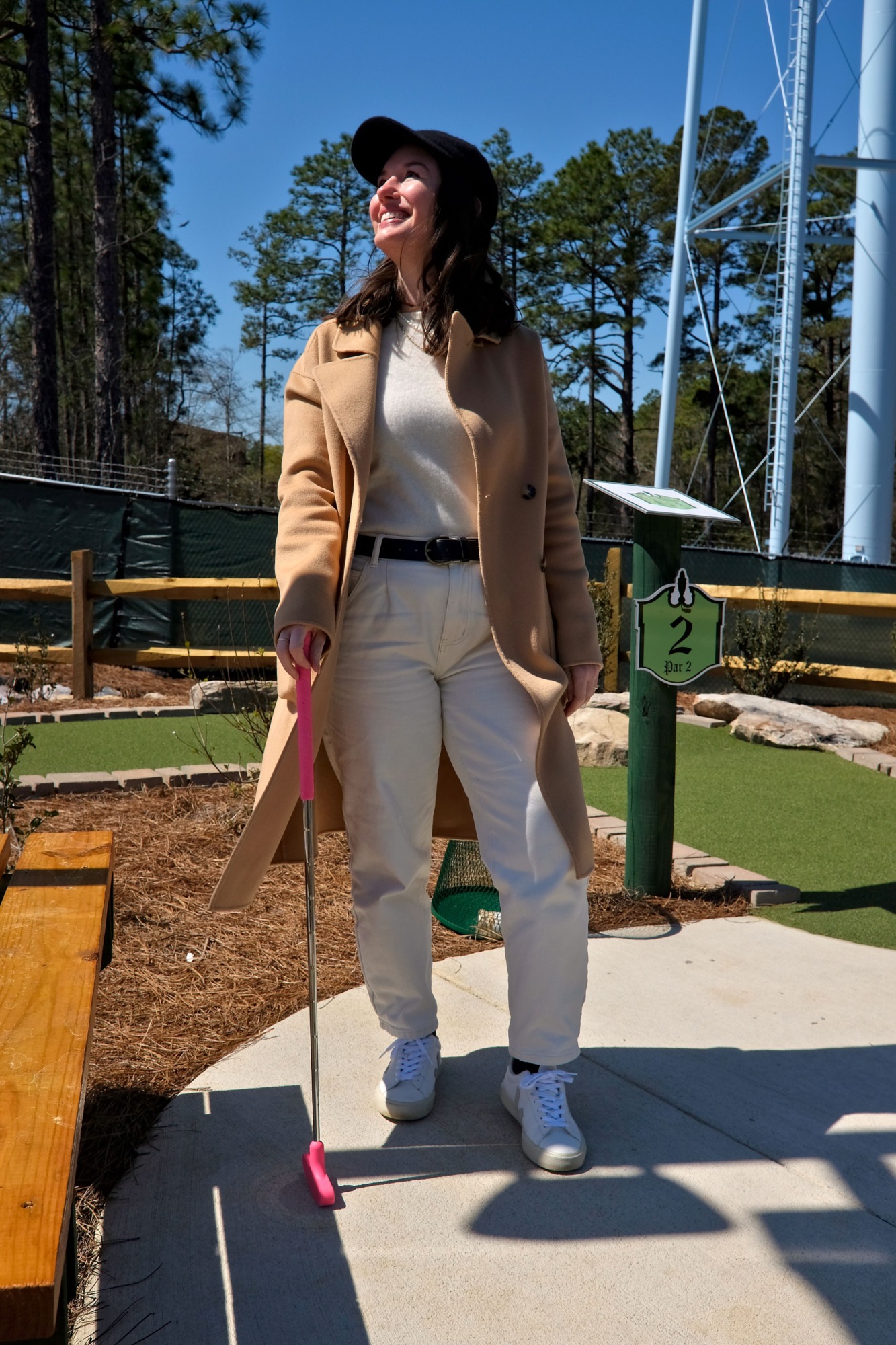Alyssa wears a white sweater with white jeans and a camel coat and a baseball cap