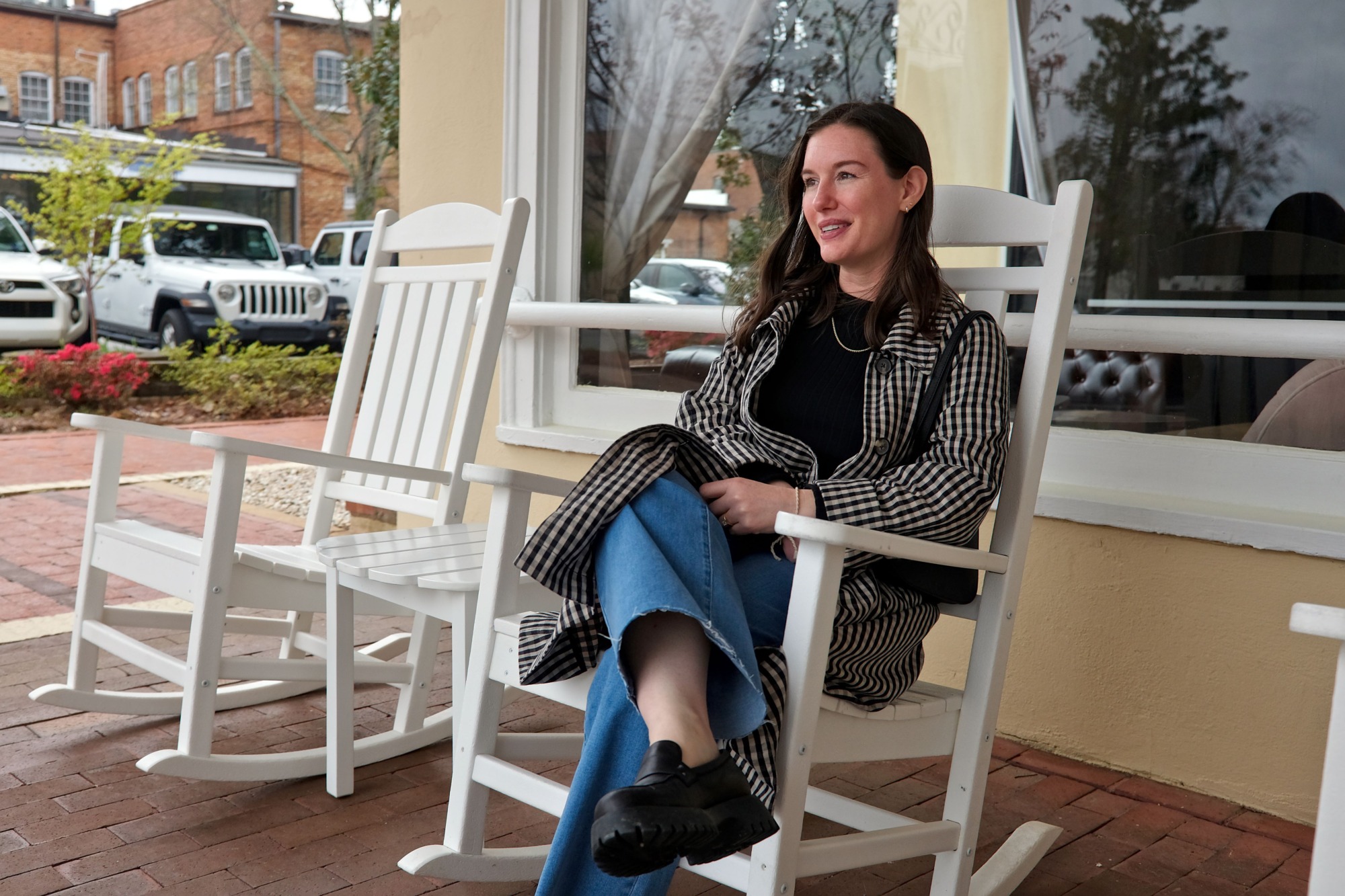 Alyssa wears a black top, blue jeans, and gingham trench and sits in a rocking chair