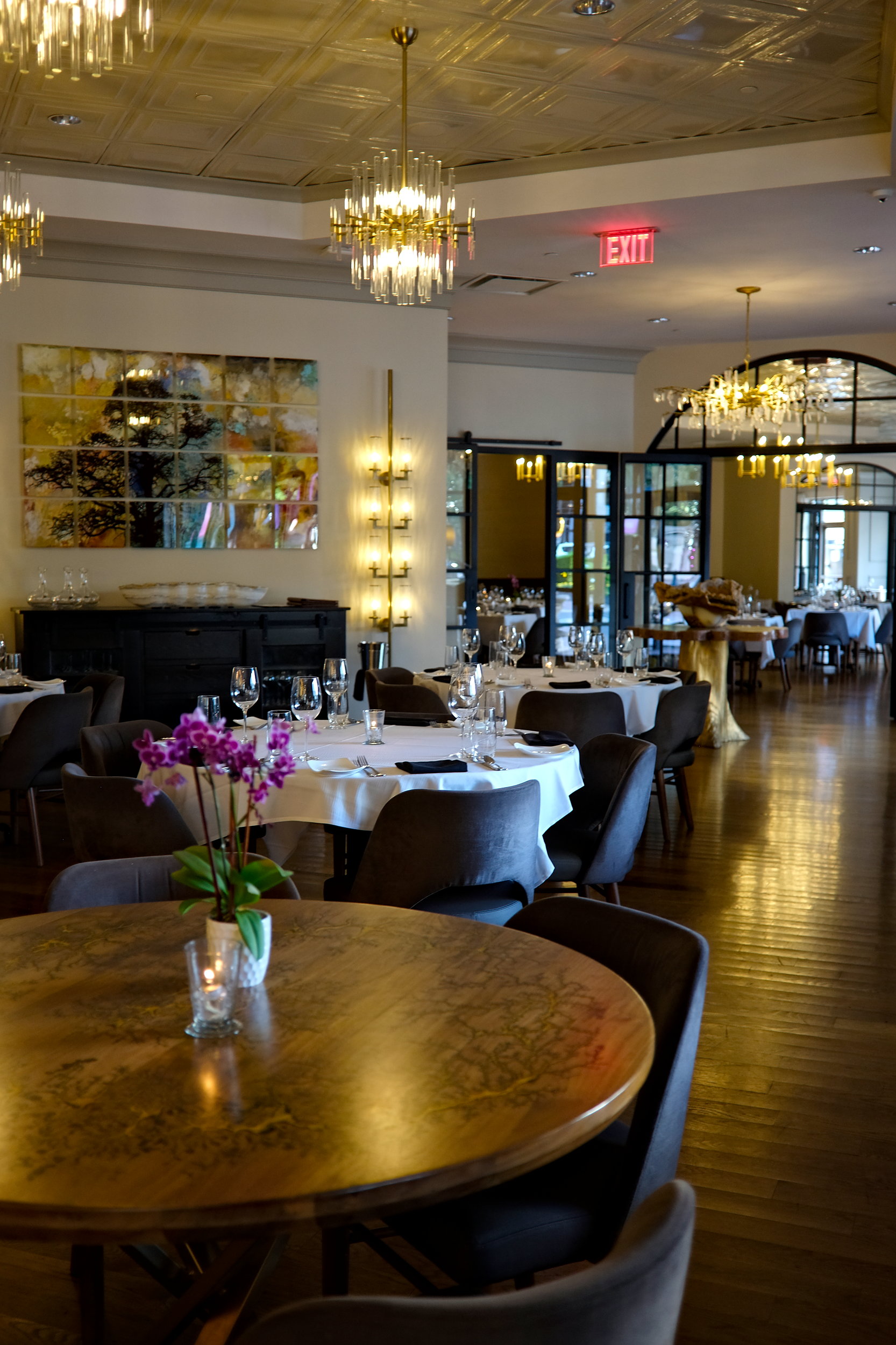 A classy dining room at Bentley’s in Charlotte with elegant chandeliers, white tablecloths, and modern art on the walls; a warmly lit, upscale atmosphere ideal for fine dining.