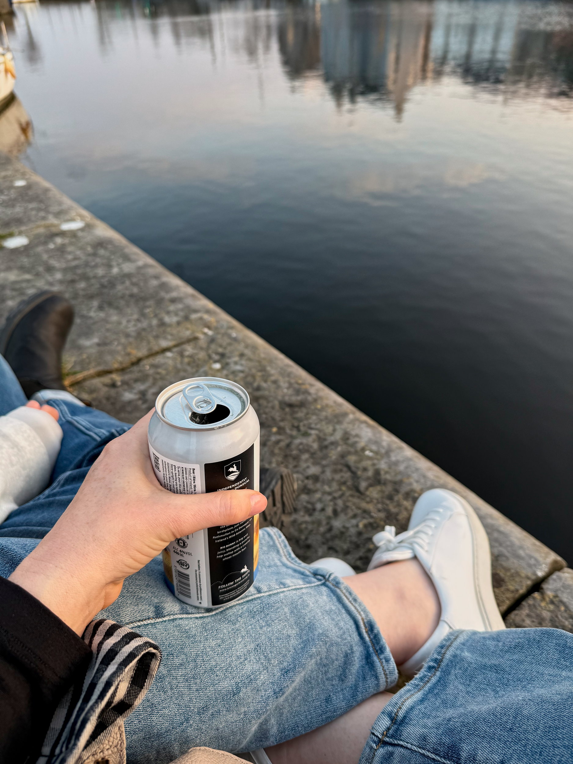 A view of Alyssa's hand holding a can of local beer as she sits along the waterway in Galway at dusk