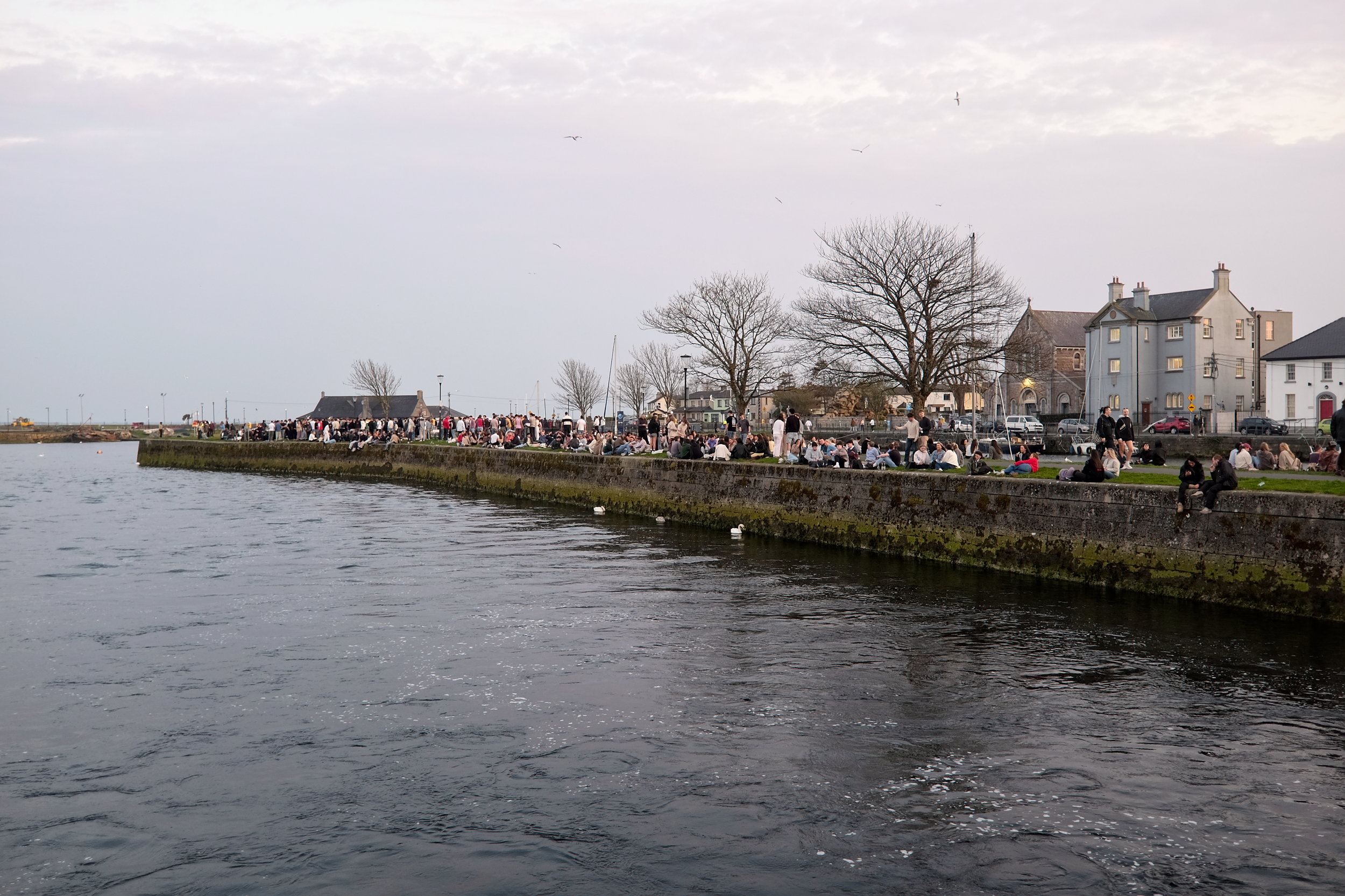 A crowd of university students sits along the waterway in Galway at dusk.