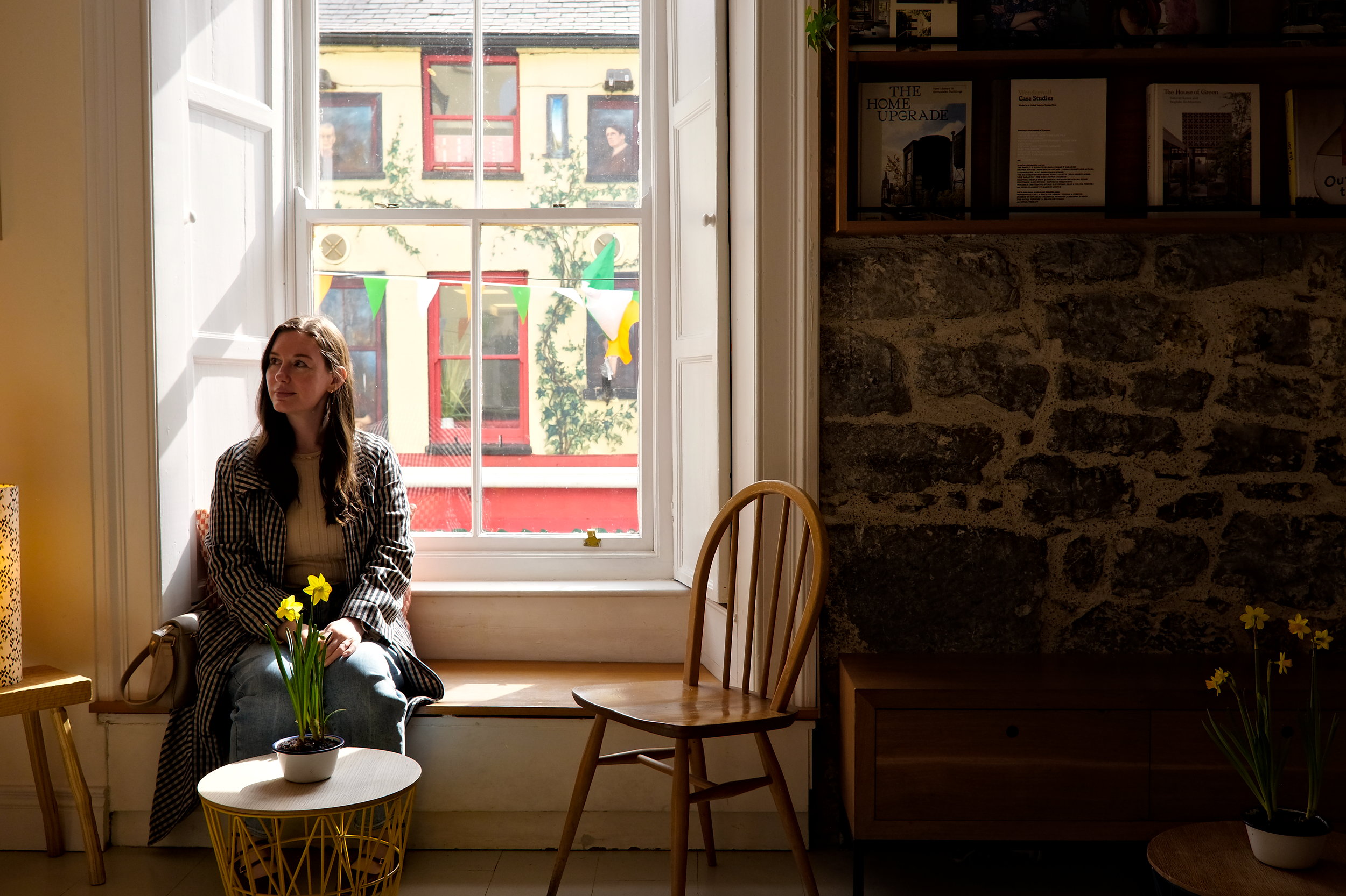 Alyssa sitting in a sunlit window seat at Coffeewerk + Press, framed by stone walls, minimal decor, and a soft street view in the background.