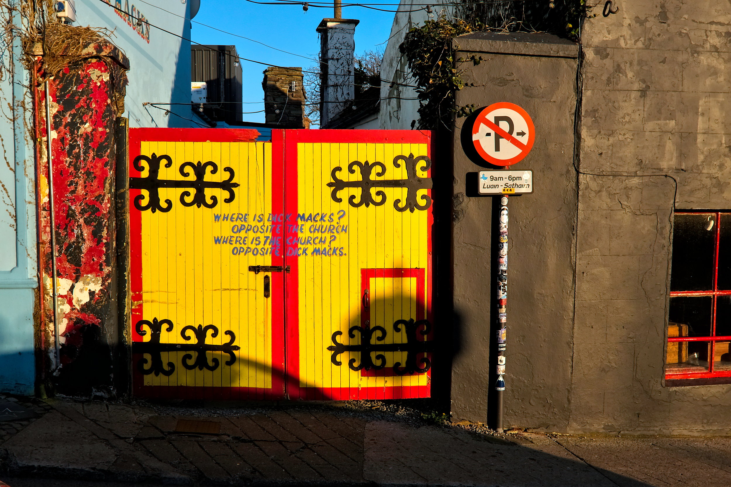 Colorful painted gate at Dick Mack’s in Dingle with a playful sign that reads, “Where is Dick Mack’s? Opposite the church. Where is the church? Opposite Dick Mack’s,” set against the backdrop of the pub’s courtyard.