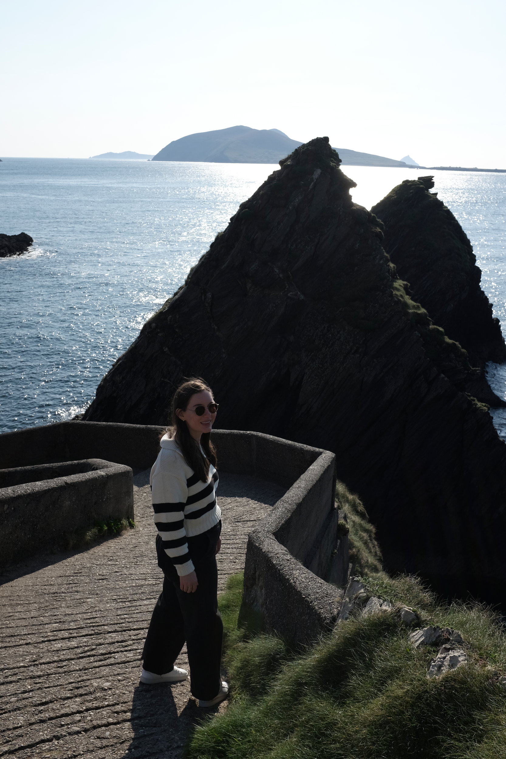 Alyssa standing partway down Dunquin Pier, surrounded by dramatic cliffs and ocean views, with the jagged path curving behind her toward the crashing waves below.