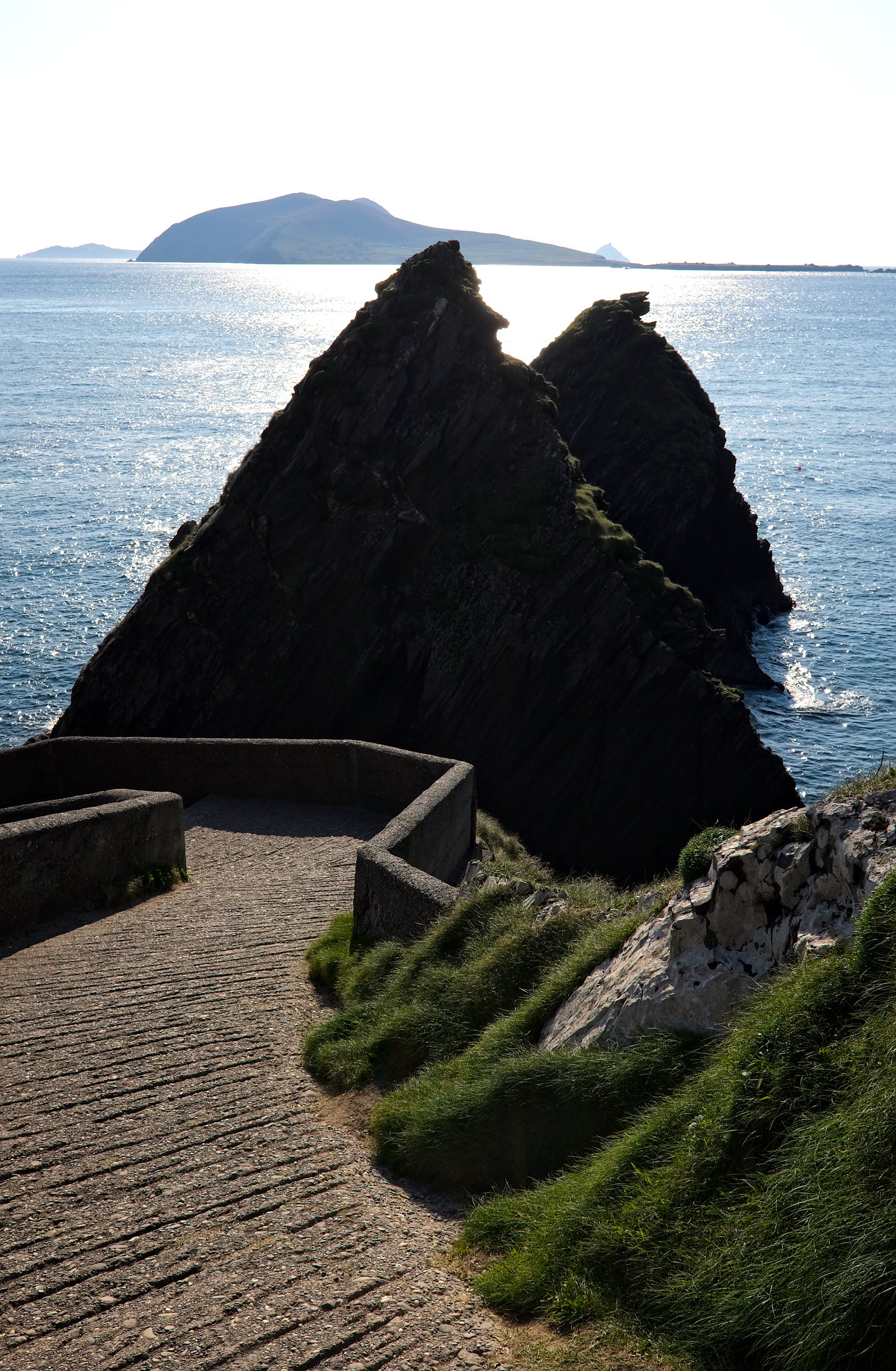 Winding concrete path of Dunquin Pier leading dramatically down steep cliffs to the ocean, with waves crashing below and the Blasket Islands in the distance.