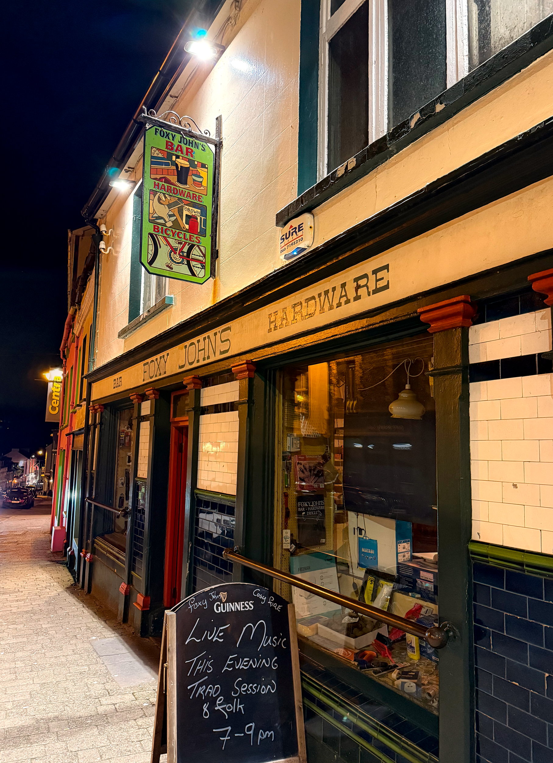 Street view of Foxy John's in Dingle, a traditional Irish pub with a rustic storefront and vintage signage that hints at its dual identity as both a bar and a hardware store.