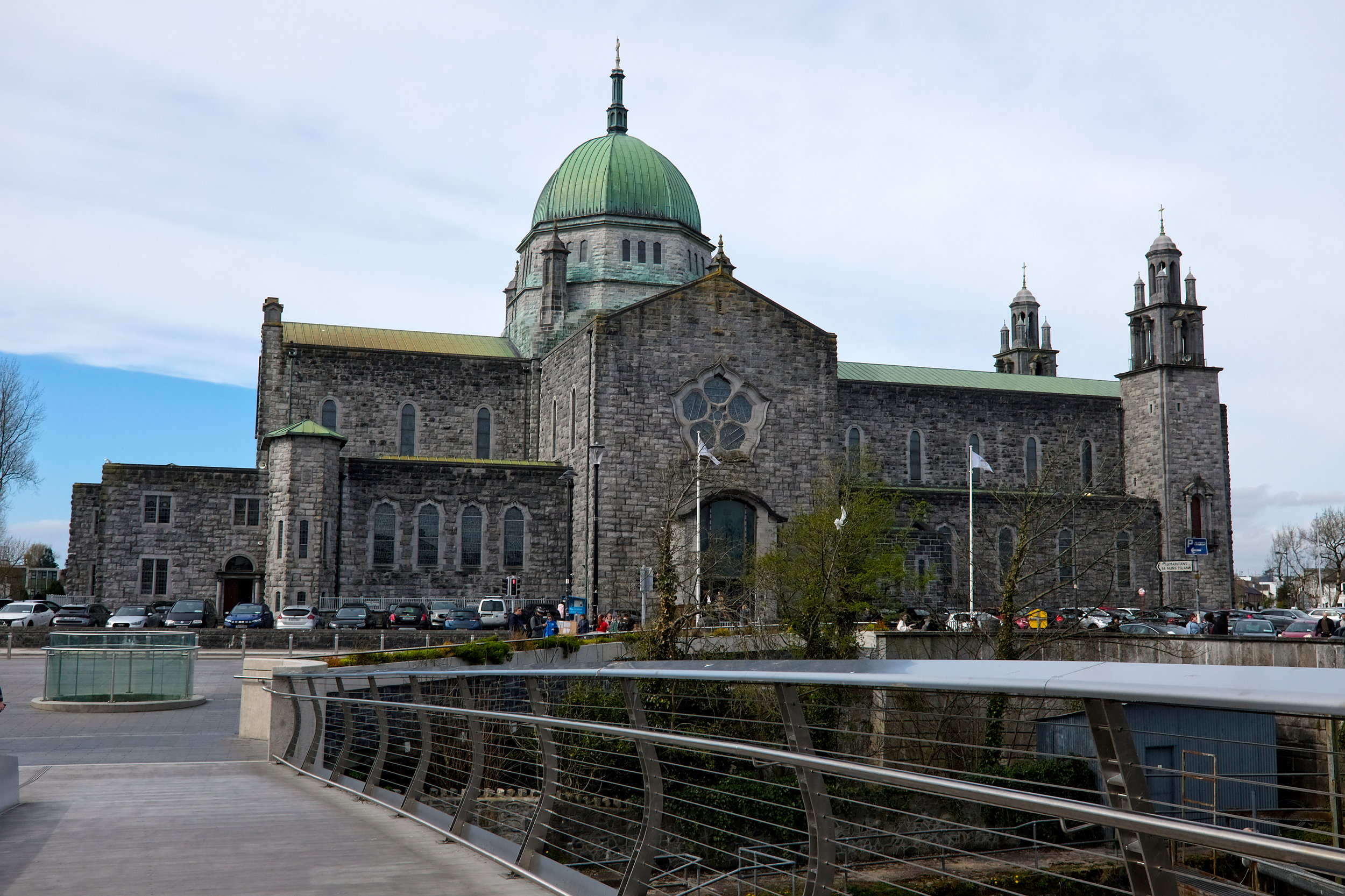 Wide-angle view of Galway Cathedral's stone façade with its large central dome, arched windows, and green copper roof, set against a partly cloudy sky and framed by lush greenery and a bridge.