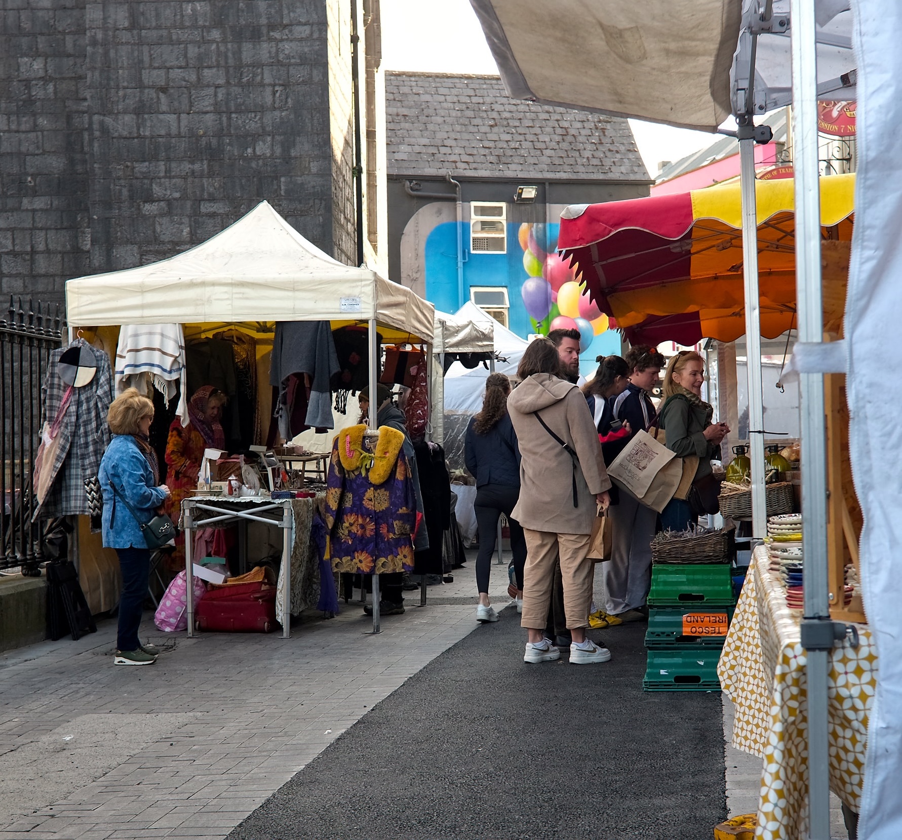 Outdoor view of Galway Market with colorful stalls and canopies, shoppers browsing local crafts and produce, and a mural of balloons on a nearby building adding a vibrant backdrop.