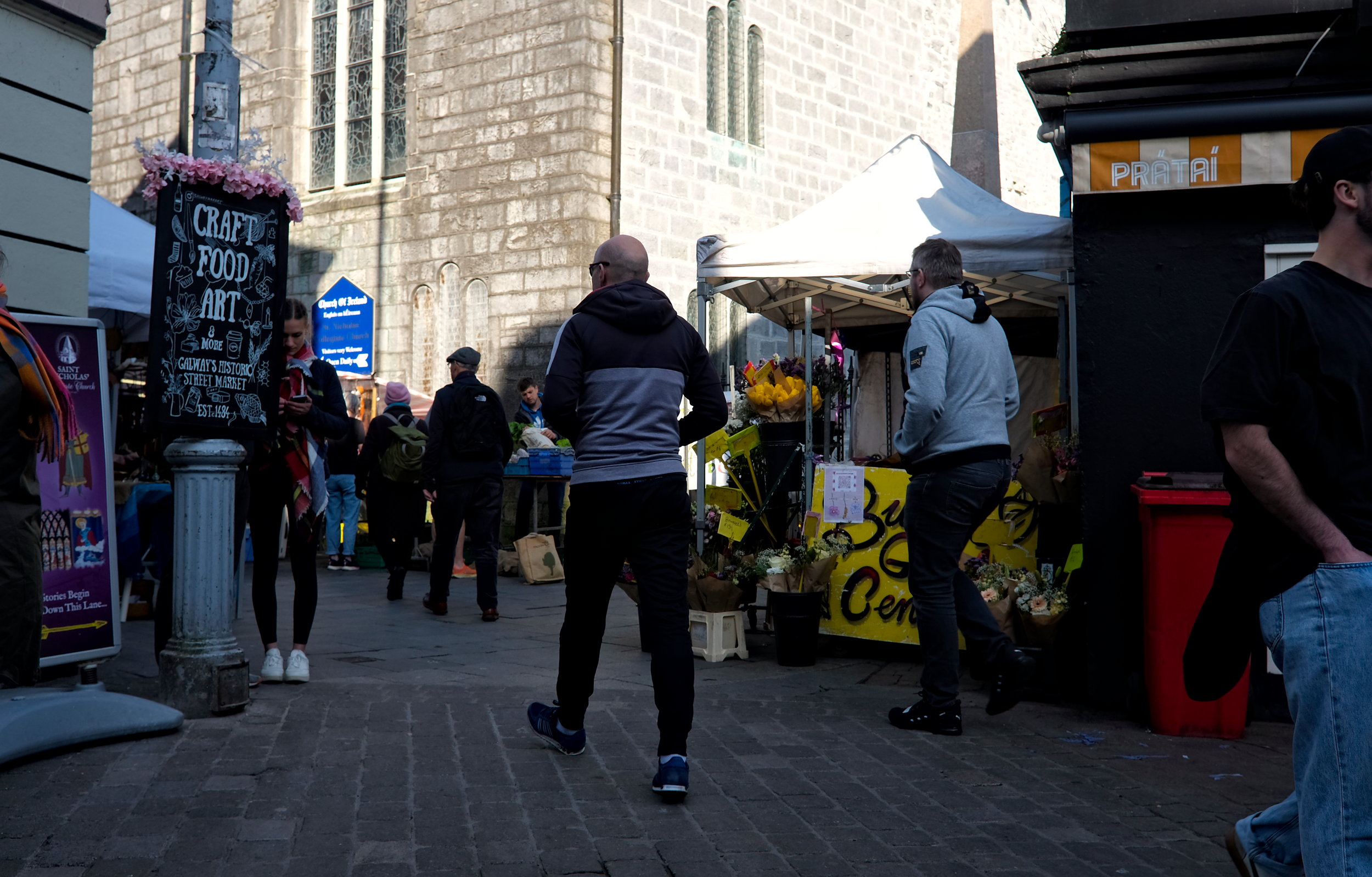 Street-level view of the entrance to Galway’s historic market, with a chalkboard sign reading “Craft, Food, Art,” people walking and browsing, and a flower stall with a bright yellow sign in the foreground.