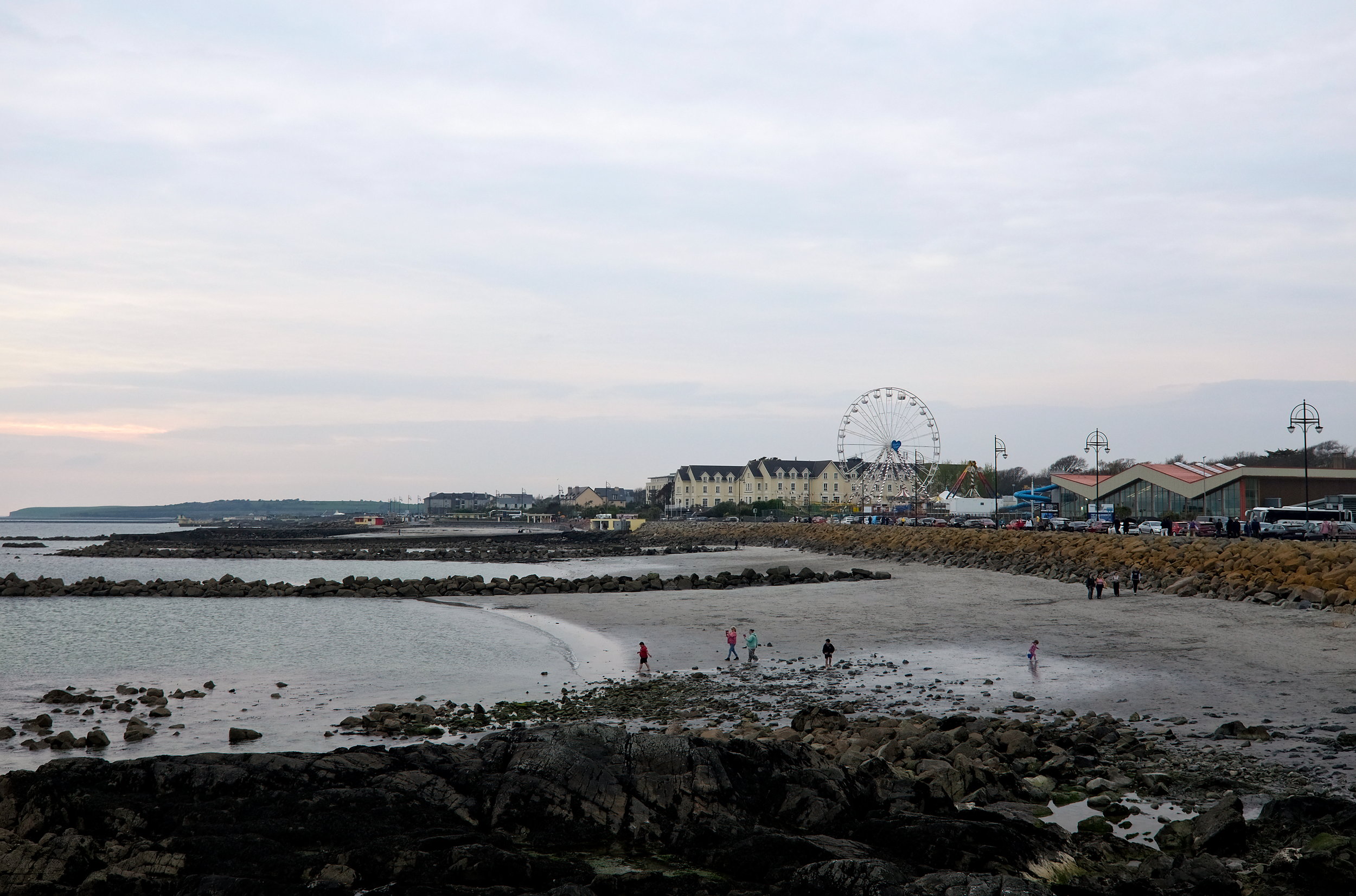 Scenic coastal view of the Atlantic Ocean along Galway’s Salthill Promenade, with a Ferris wheel rising in the distance, waves breaking gently along the shore, and walkers dotting the path.