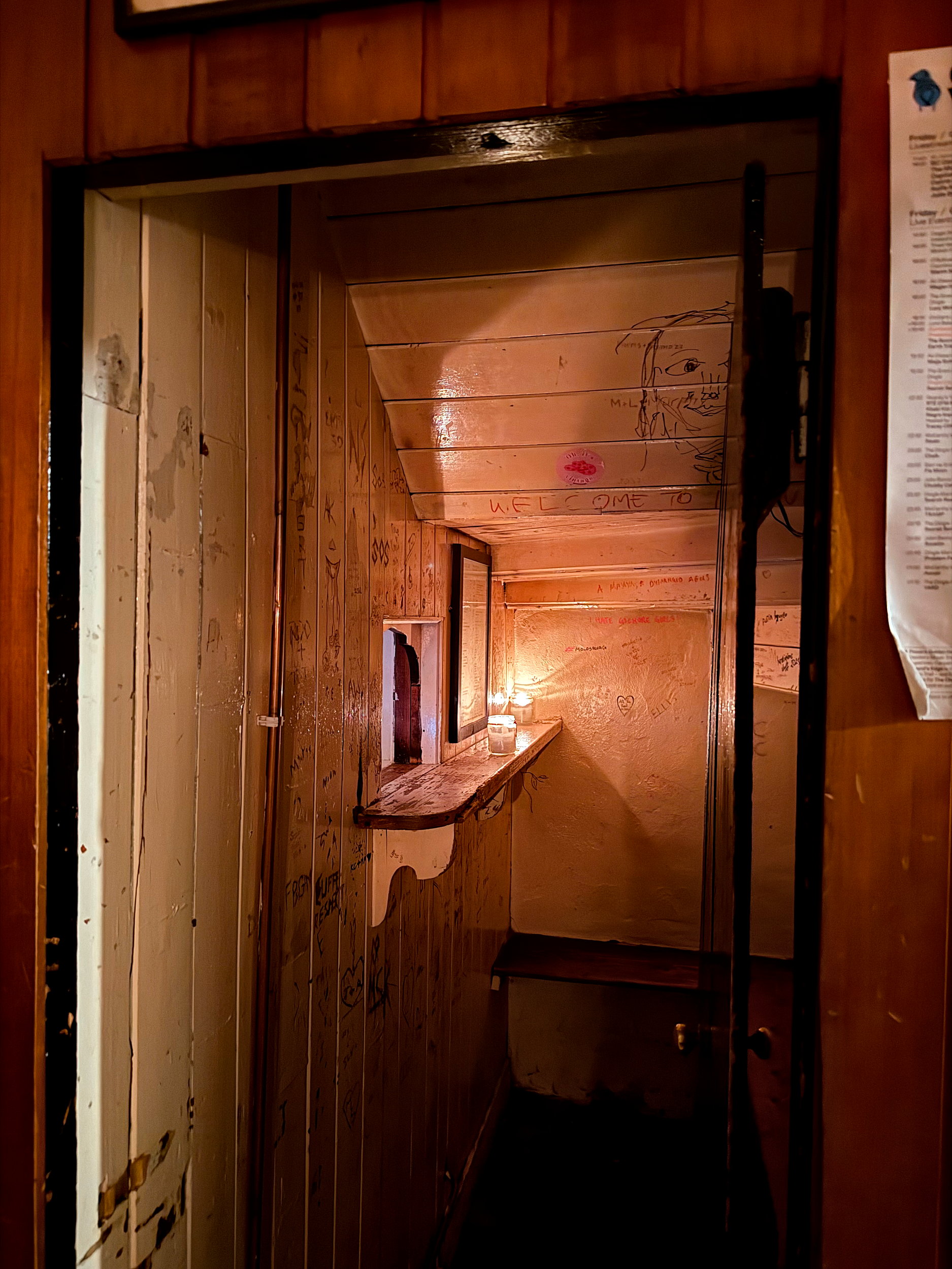 Narrow, dimly lit wooden booth inside Kennedy's Bar with candlelight glowing on a small ledge, graffiti-covered walls, and a cozy, rustic atmosphere.