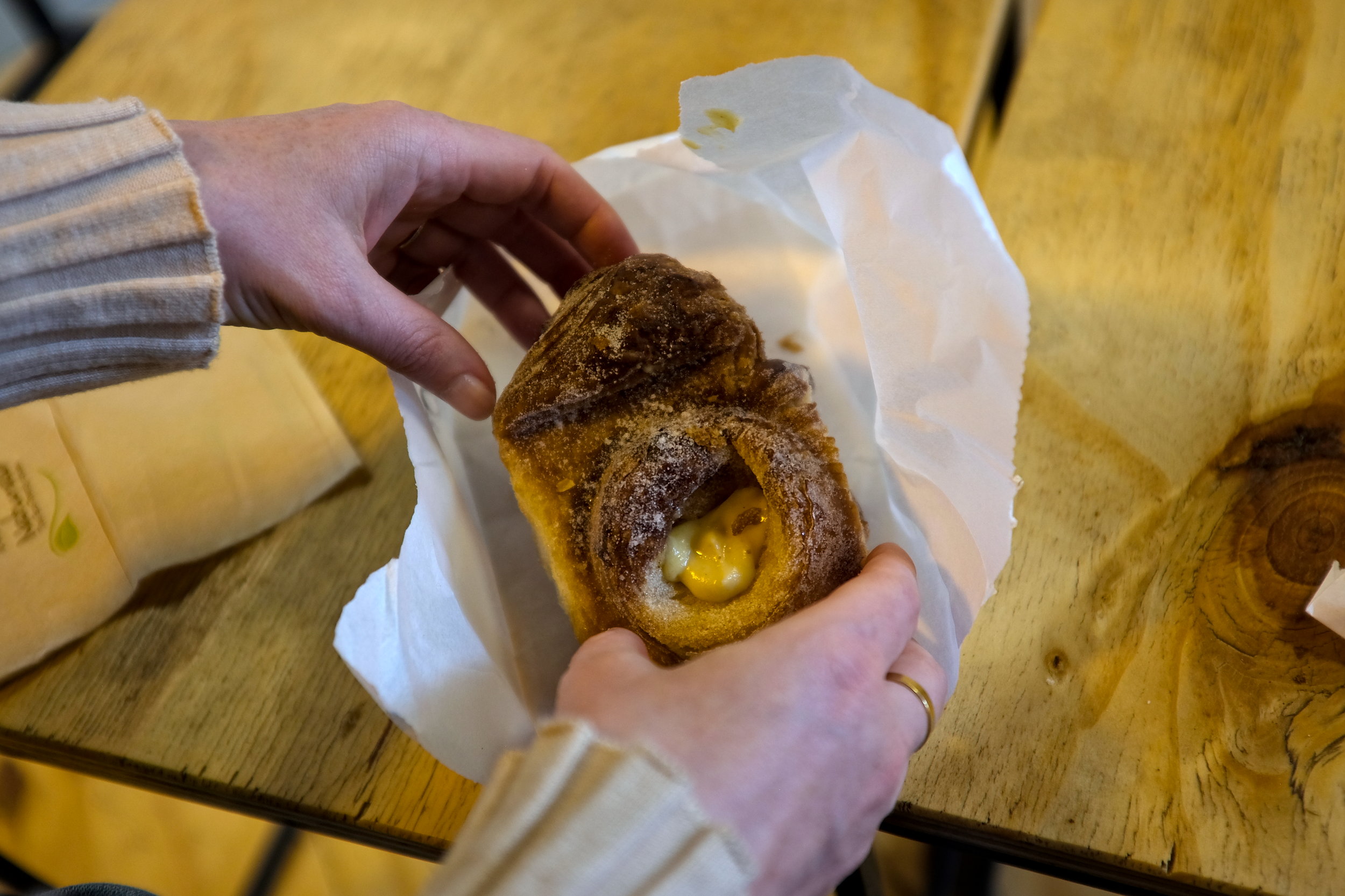 Close-up of two hands gently holding a passionfruit croissant, revealing its airy layers and vibrant yellow filling, with pastry flakes scattered on a white paper and table beneath.