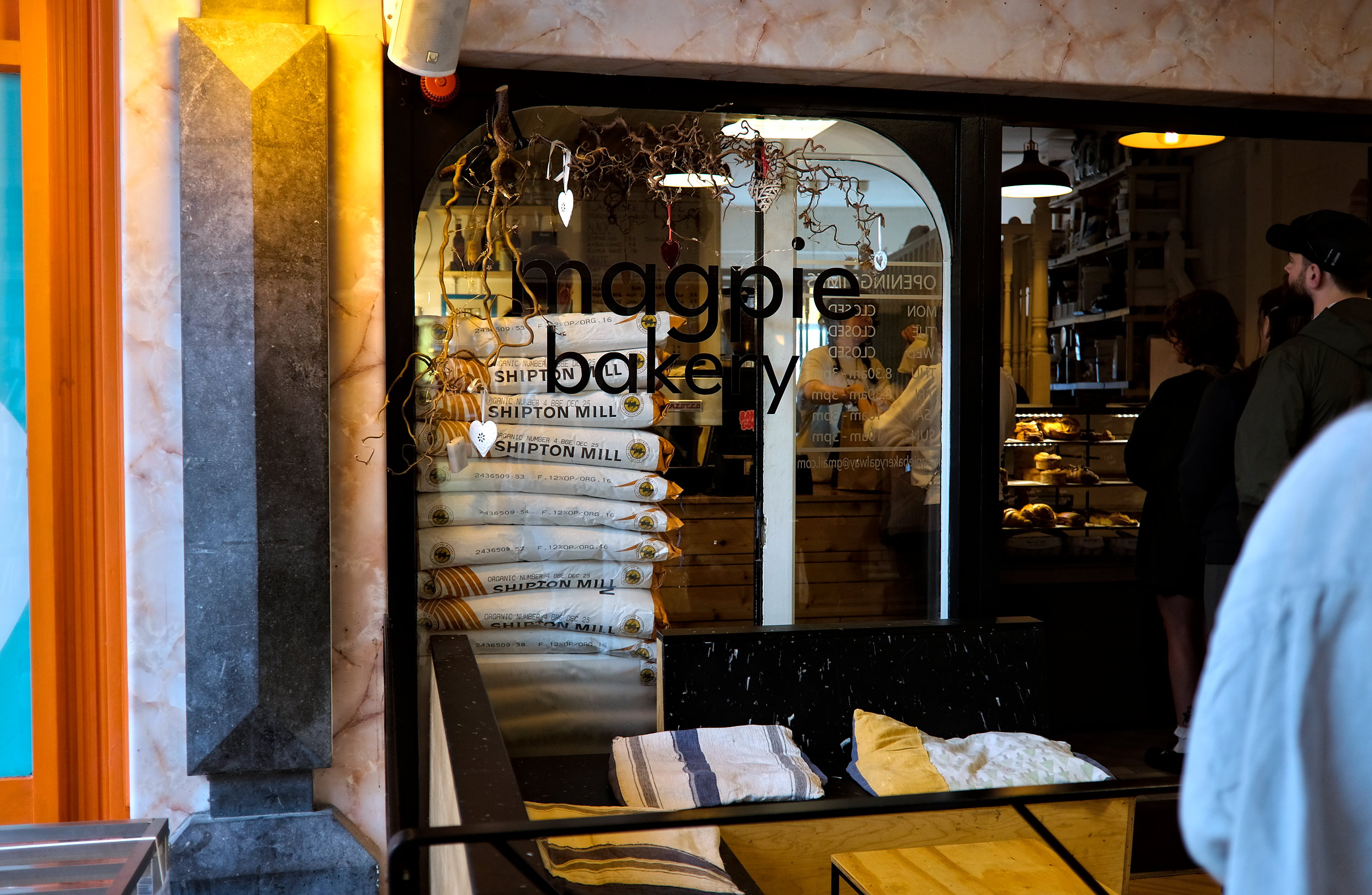 Exterior view of Magpie Bakery's entrance with a line of customers waiting outside, set against a charming storefront with large windows and a simple, modern sign.