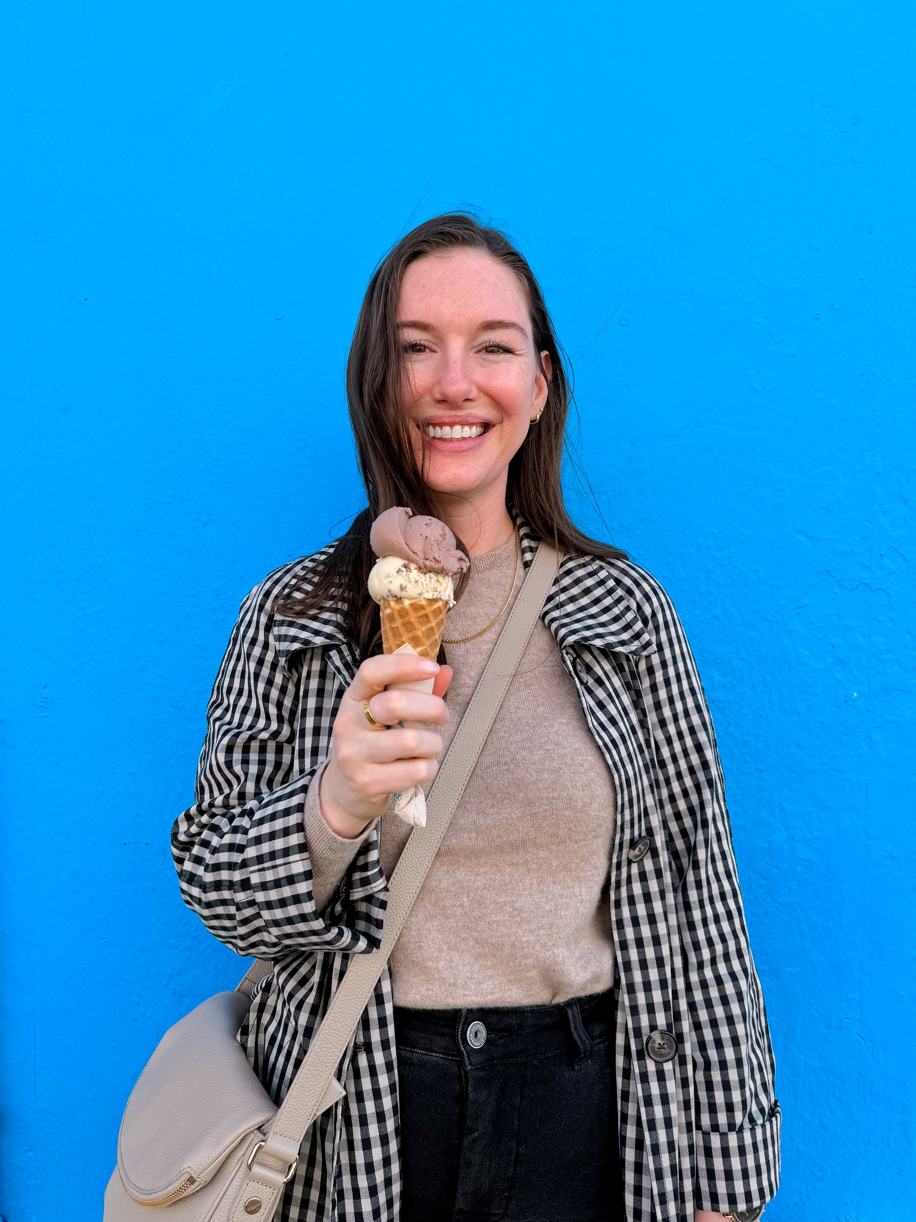 Alyssa smiles while standing in front of a bright blue wall holding a double-scoop ice cream cone with chocolate whiskey and Irish brown bread flavors. She wears a black-and-white checkered coat, beige sweater, and crossbody bag.