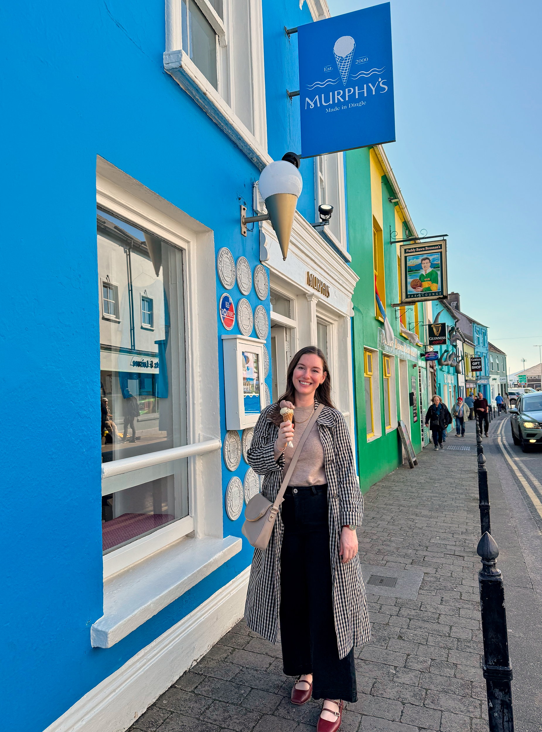 Alyssa enjoying an ice cream cone while walking down a vibrant street in Dingle, surrounded by brightly painted shopfronts and a cheerful, relaxed atmosphere.