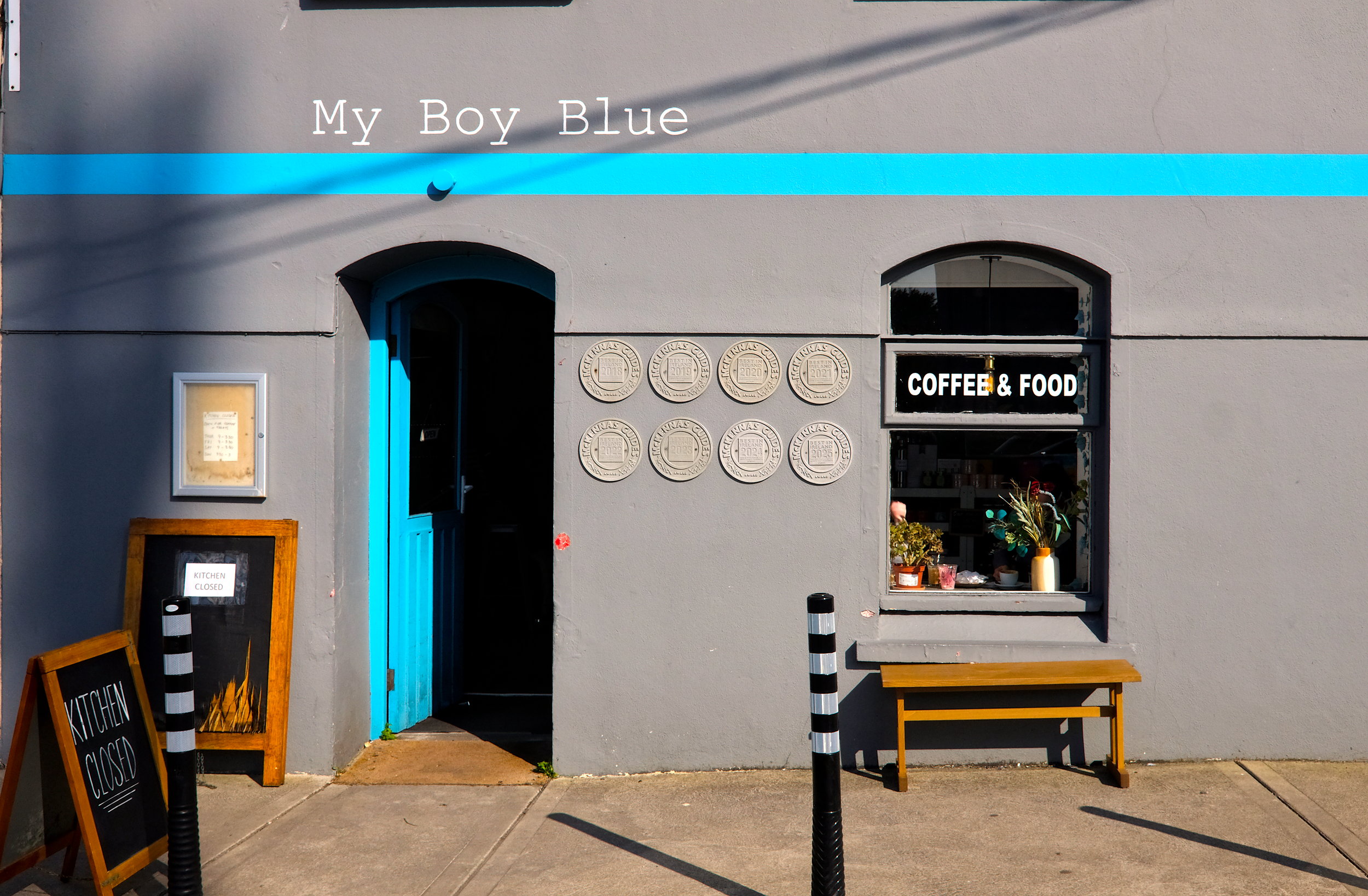 Exterior of My Boy Blue café in Dingle with a grey and blue facade, a black-and-white "Coffee & Food" sign in the window, and multiple award plaques on the wall. A sandwich board reads "Kitchen Closed," and a yellow bench sits beneath the window.