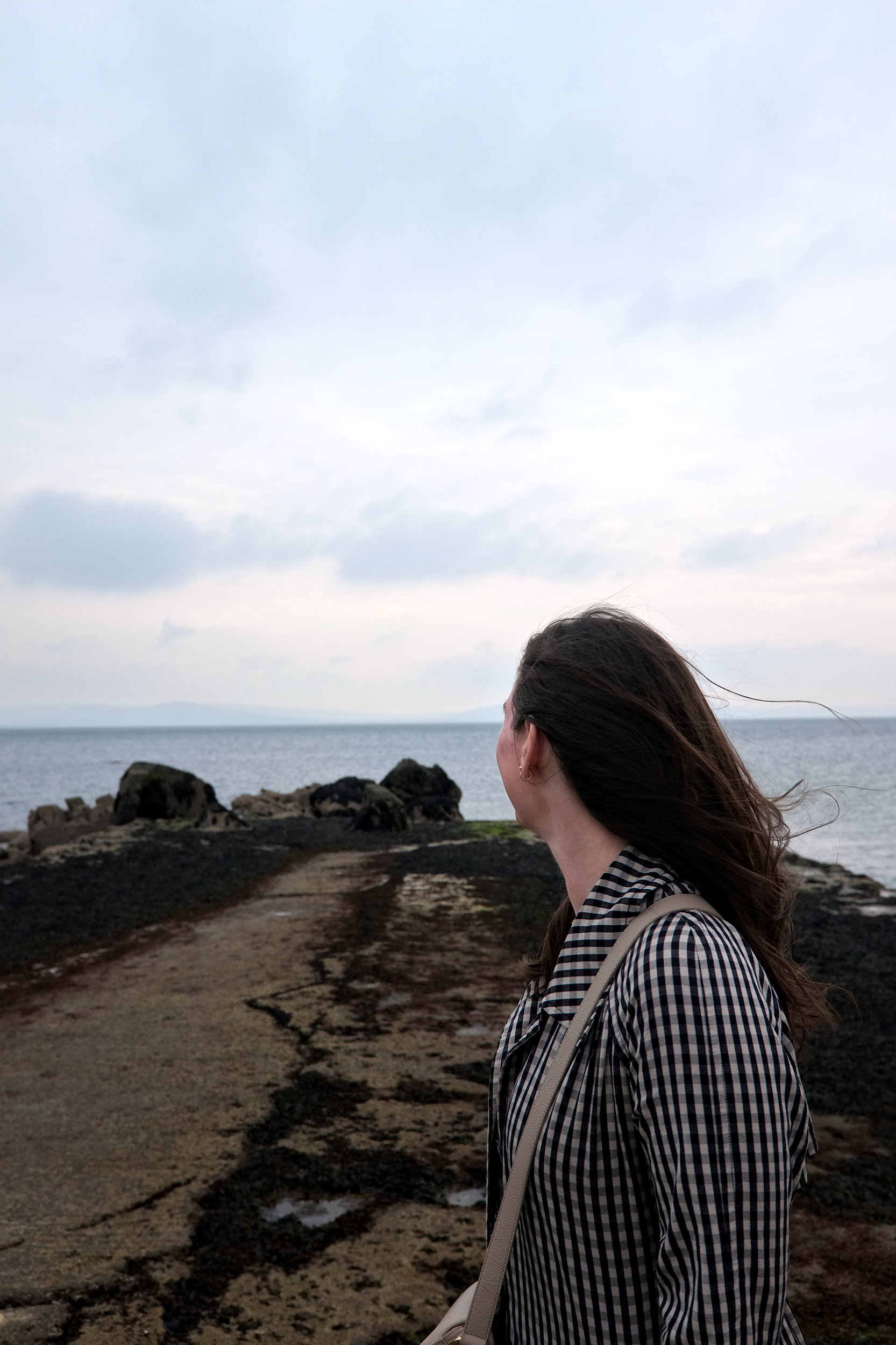 Candid shot of Alyssa walking along Salthill Promenade, gazing out at the Atlantic Ocean, with wind in her hair and a calm, expansive seascape stretching beyond her.