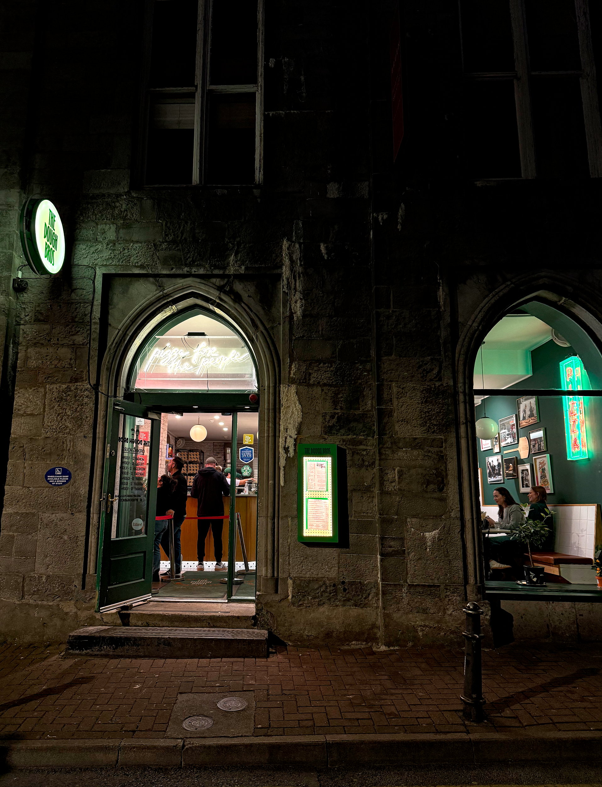 Nighttime view of The Dough Bros restaurant exterior, warmly lit with a glowing sign above the entrance and people gathered inside, creating a cozy, inviting late-night vibe.