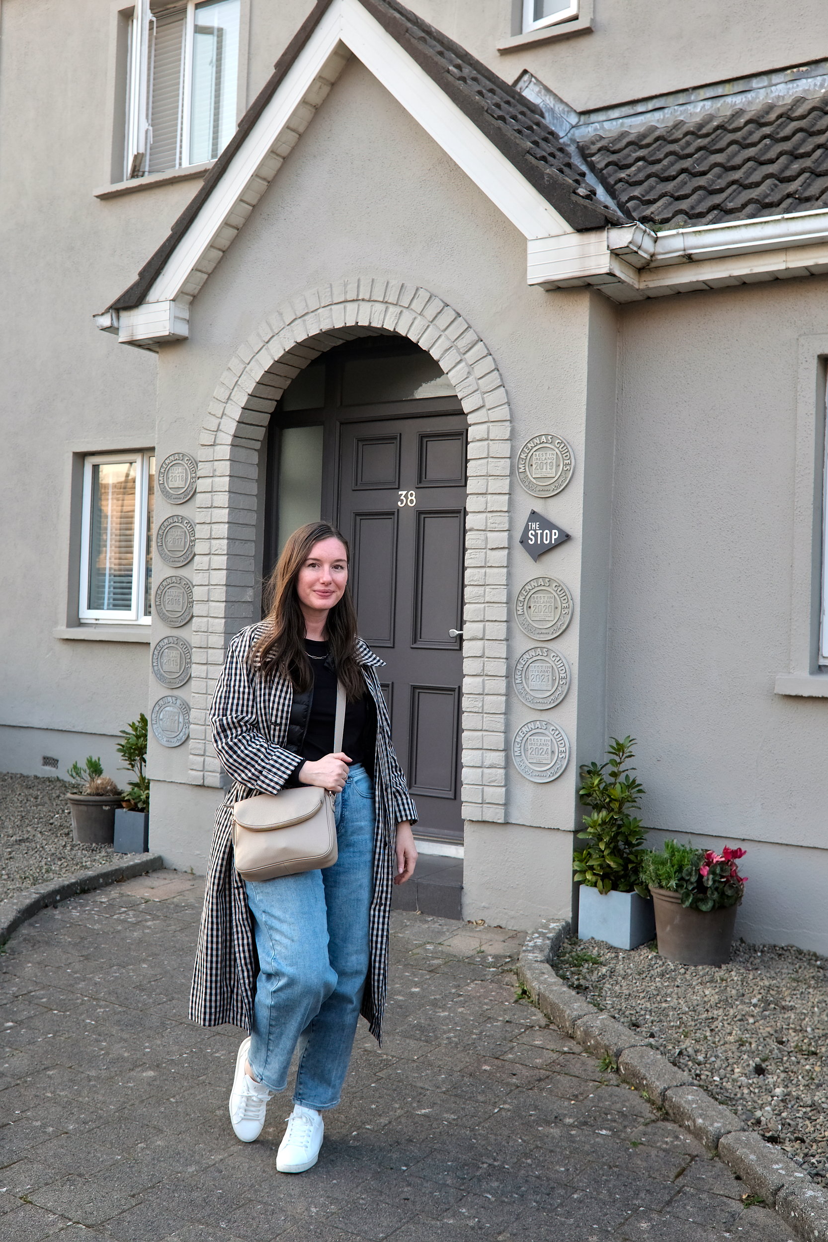 The author stands outside the entrance of The Stop Galway, a boutique bed and breakfast with a grey facade, arched brick doorway, and the number 38 above the door. Decorative award plaques and potted plants frame the stylish entryway.