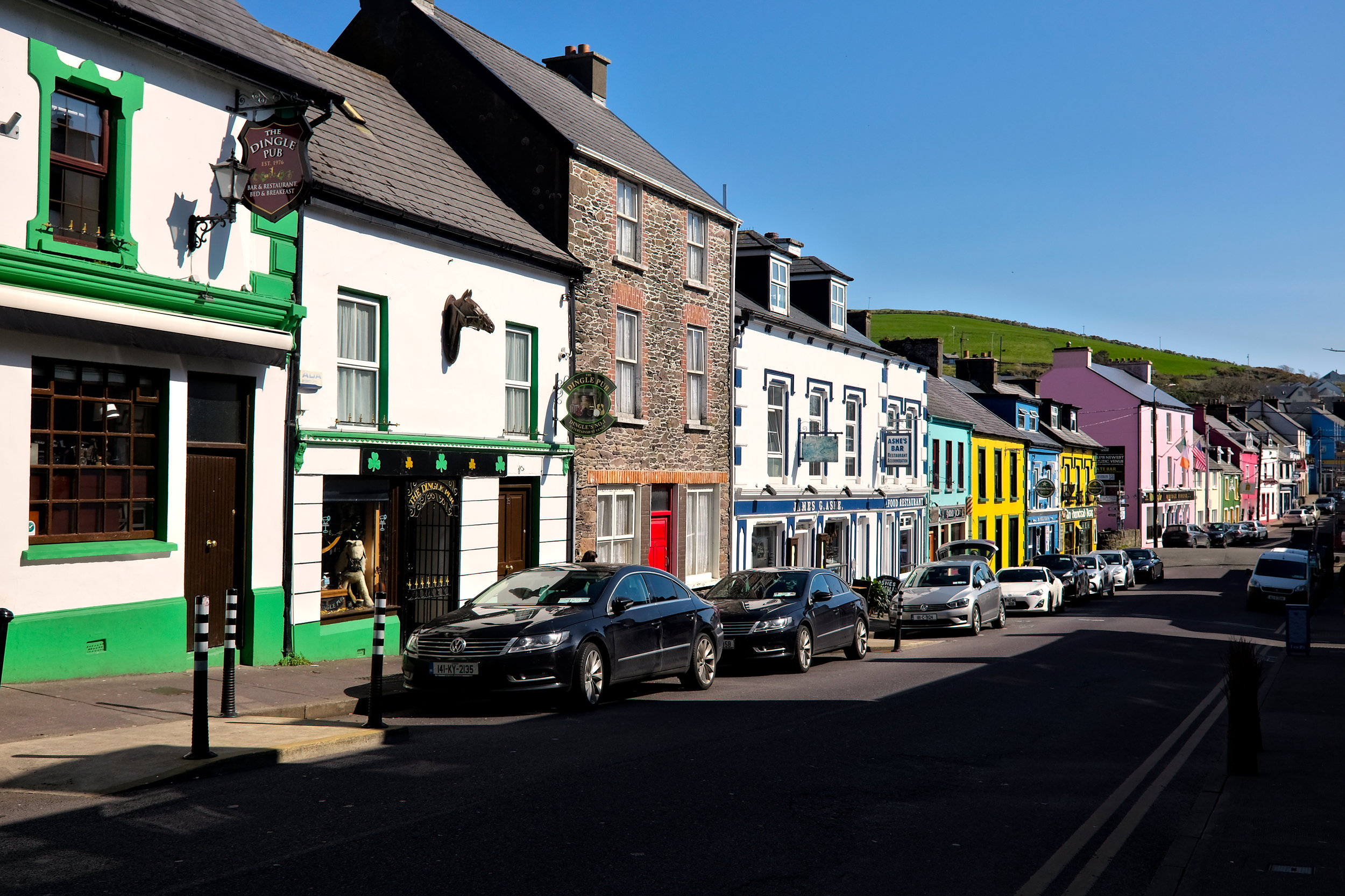 Vibrant street scene in Dingle lined with colorful shopfronts in shades of purple, blue, yellow, and green, with a lively, small-town charm in the air.