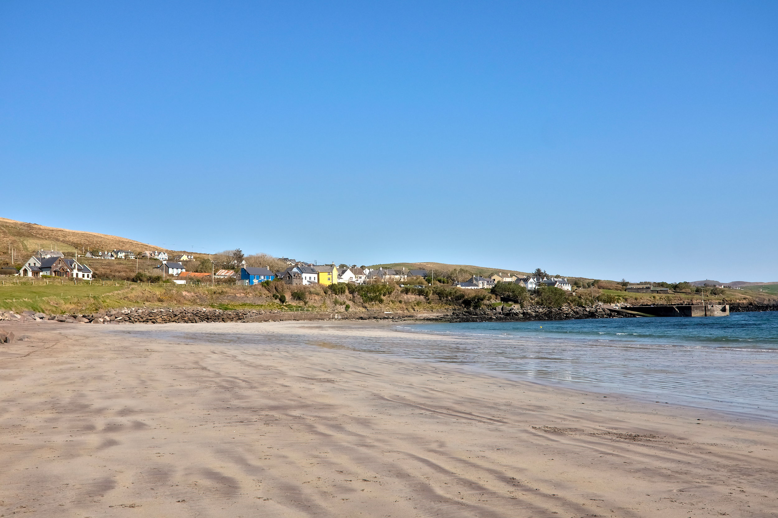 Wide stretch of sandy Ventry Beach with gentle waves lapping the shore, backed by grassy dunes, cute homes, and distant hills under an open sky.
