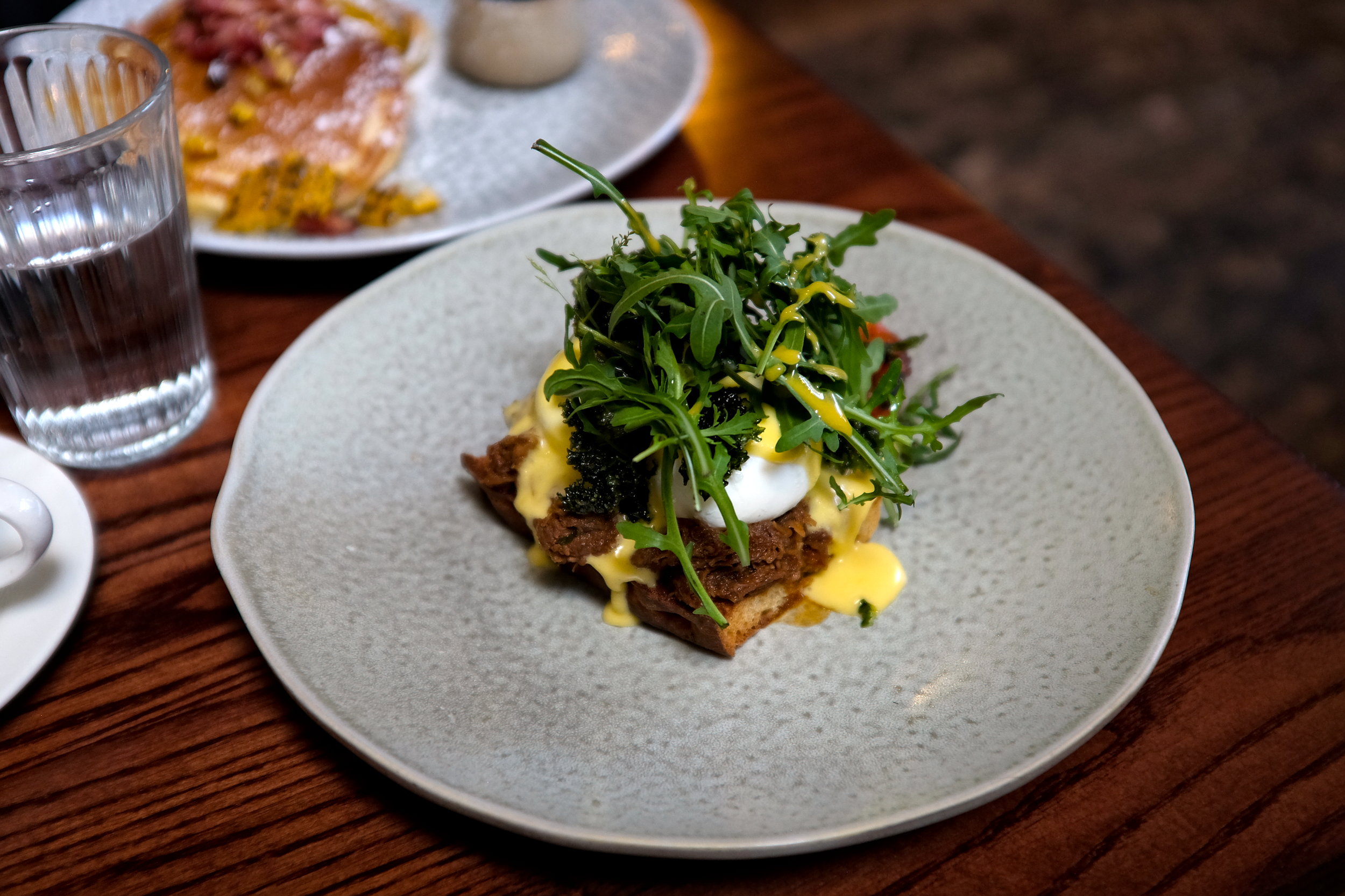 A grey plate with a pork benedict, including focaccia, pork, poached egg, tajin, and arugula, all topped with a drizzled yellow sauce