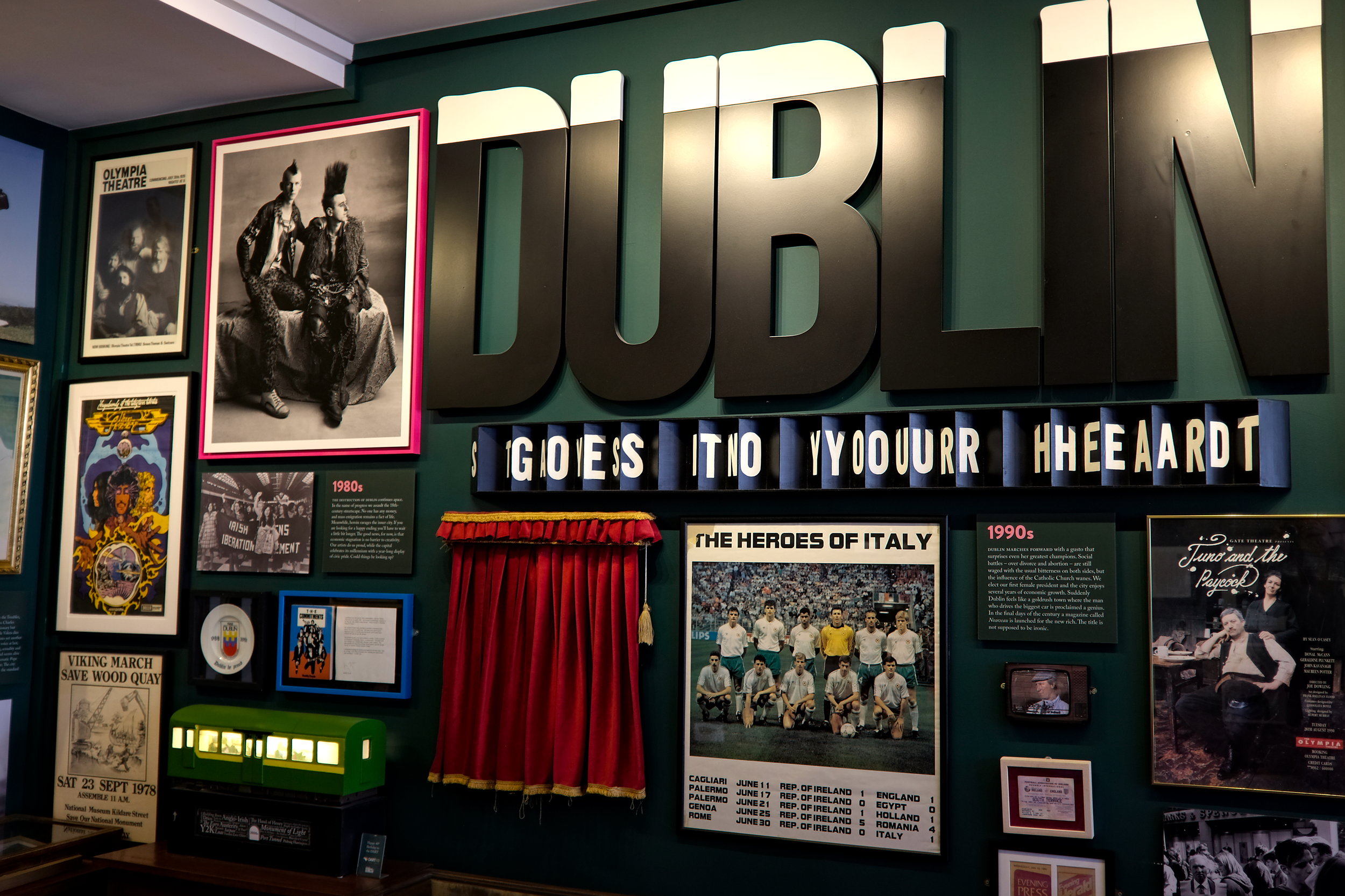 A vibrant museum wall display with a large “DUBLIN” sign and various framed photos, posters, and memorabilia, including a punk rock portrait, a red curtain, a poster titled “The Heroes of Italy,” and text reading “IT GOES INTO YOUR HEART” in block letters.