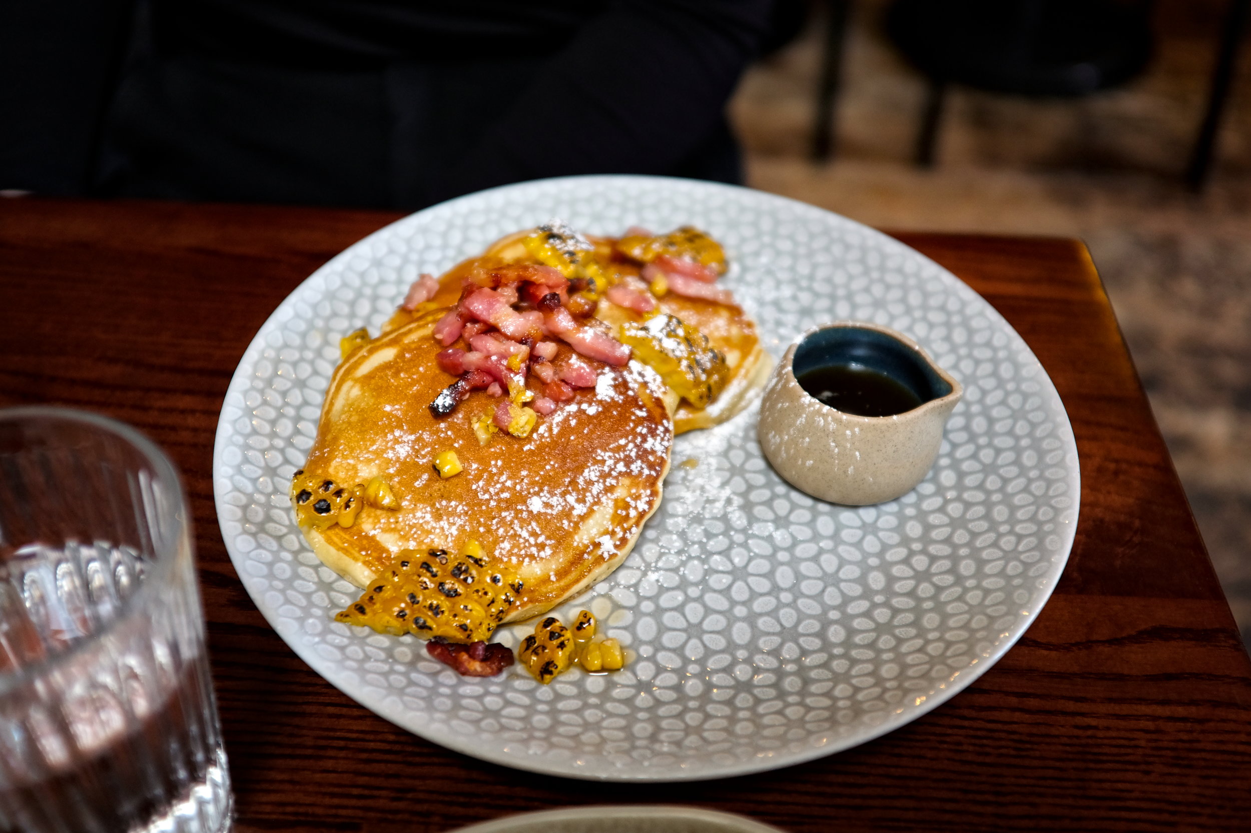 A plate of three pancakes topped with grilled corn and lardons of bacon, with a side of maple syrup