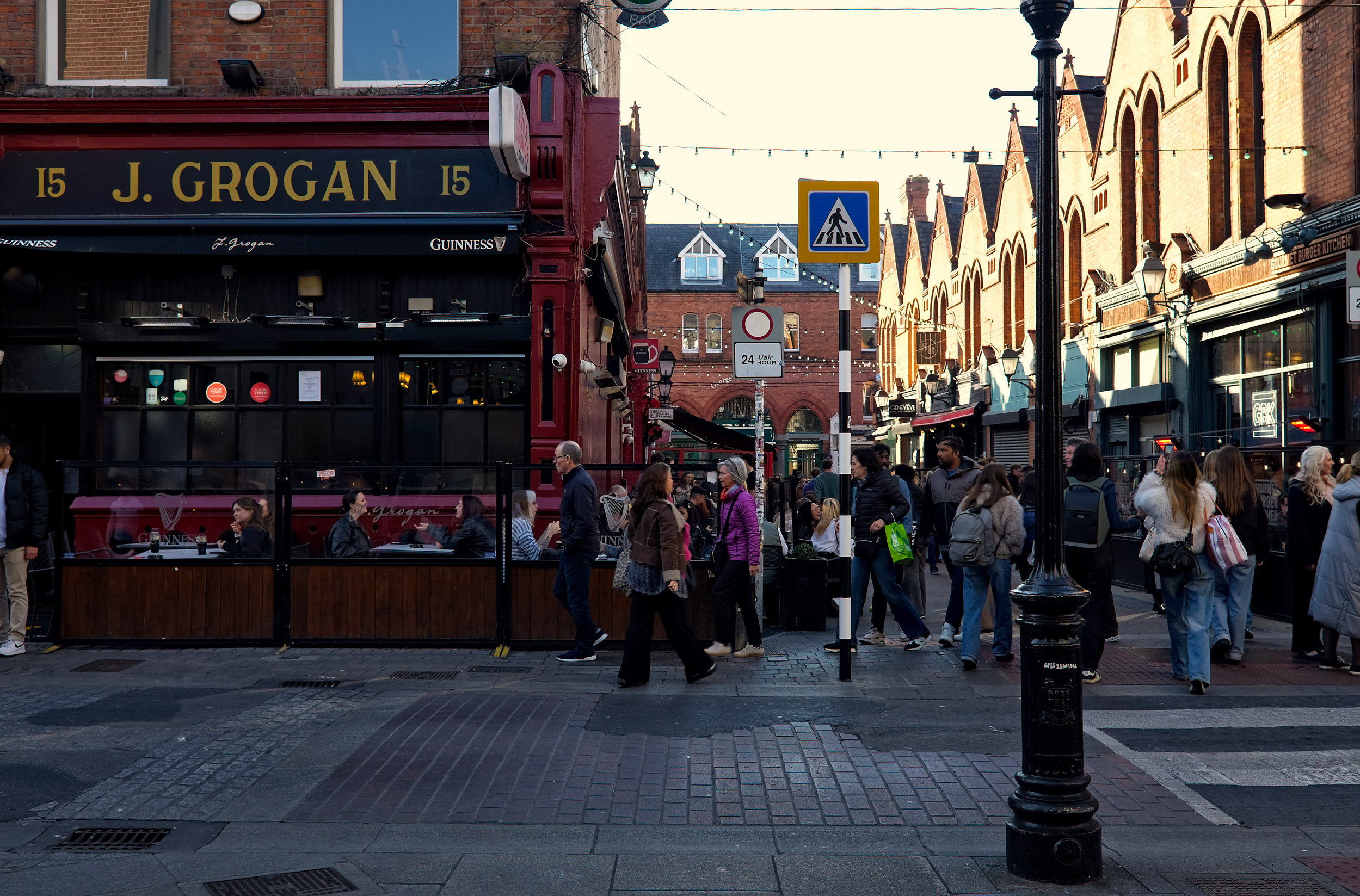 A bustling street scene outside Grogan’s pub in Dublin, featuring people walking past and sitting at outdoor tables. The pub has a classic black and red exterior with “J. Grogan” signage, and the street is lined with red-brick buildings and hanging lights.