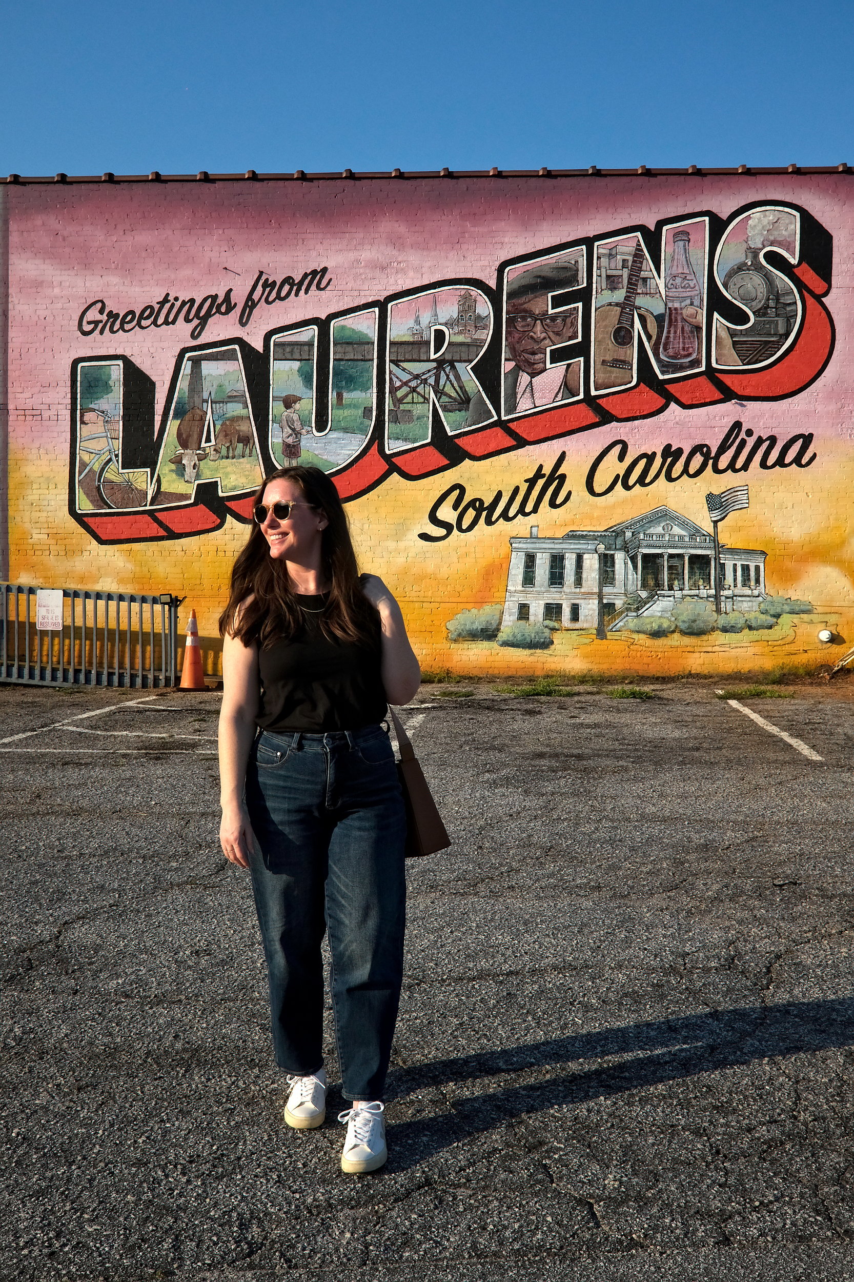 A woman in sunglasses, a black tank top, and high-waisted jeans smiles while walking in front of a colorful mural that reads “Greetings from Laurens, South Carolina.” Each letter in "Laurens" contains illustrated scenes, including a Coca-Cola bottle, historic buildings, and people, representing the local culture and history.