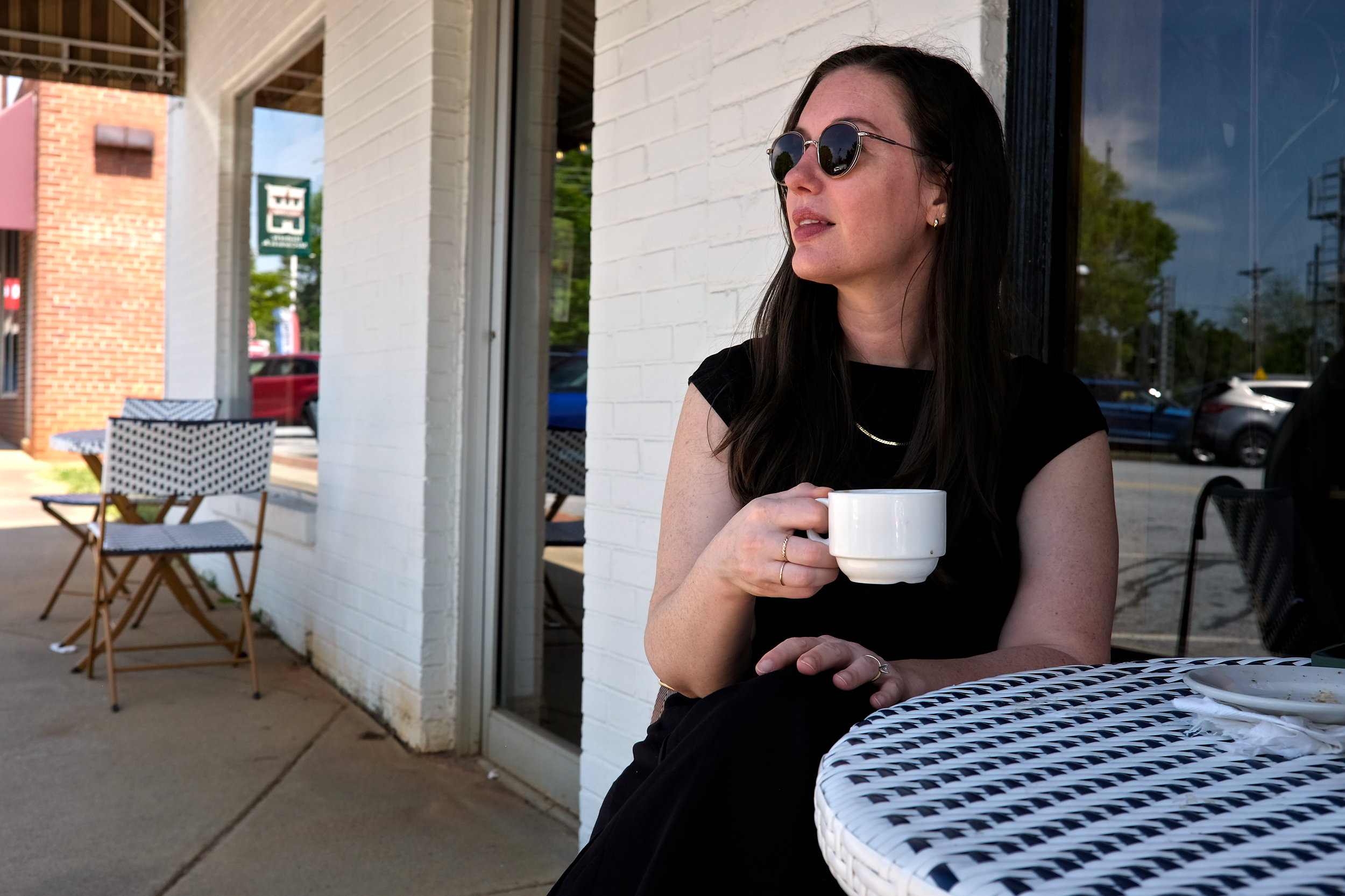 A woman wearing sunglasses and a black outfit sits at a white café table outdoors in McCormick, holding a white coffee cup. She gazes off to the side with a relaxed expression, with a mostly empty plate and napkin beside her; black-and-white patterned chairs and a brick building are visible in the background.