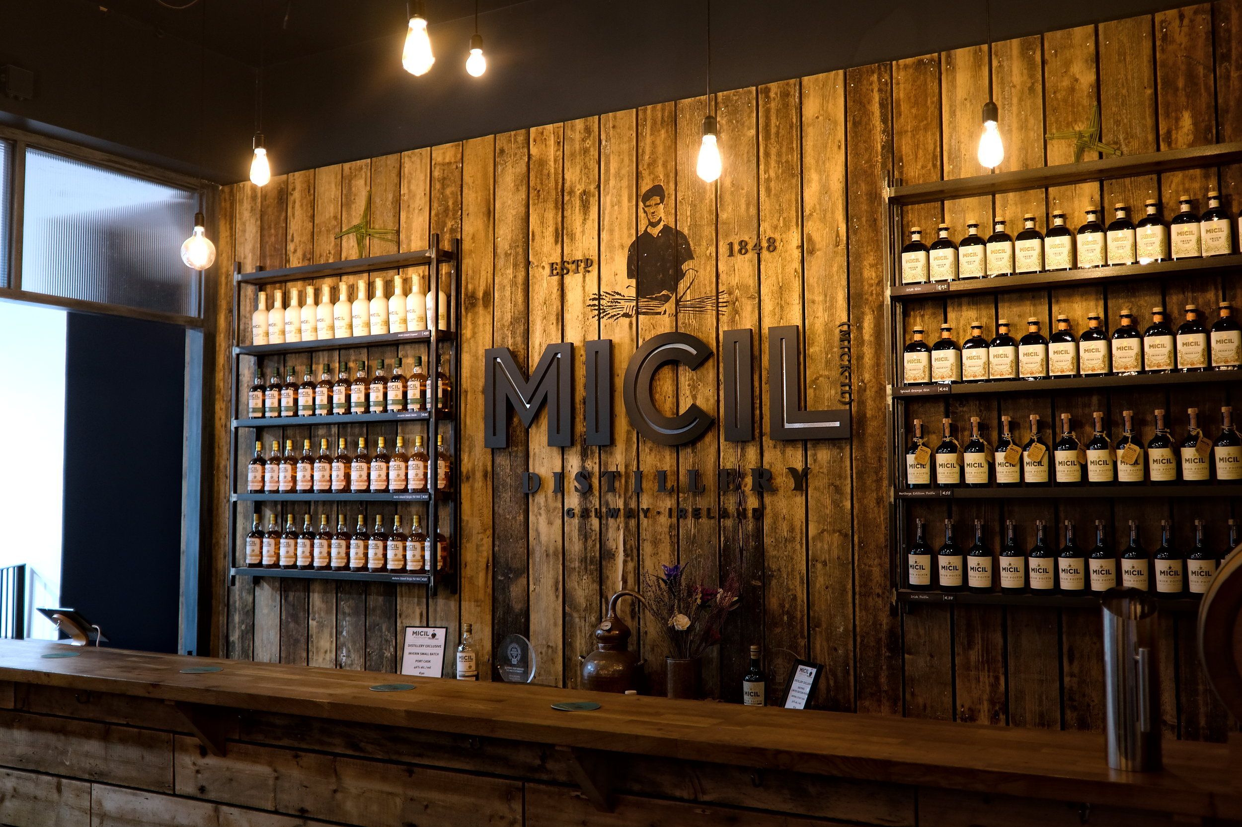 Display wall at Micil Distillery featuring the Micil logo in bold letters above shelves lined with neatly arranged bottles of whiskey, poitín, and other spirits.
