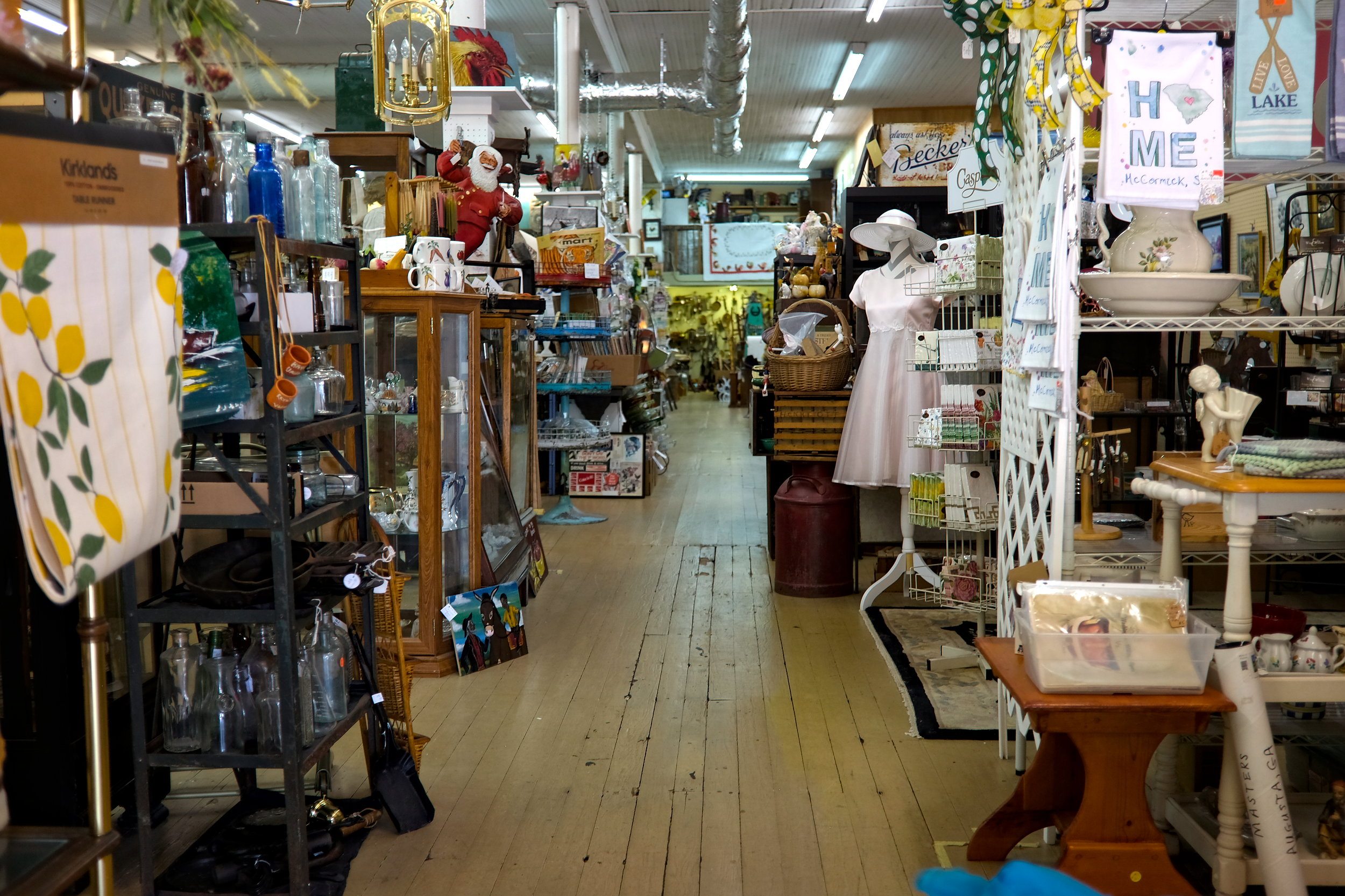 Storeys at The Red Rooster is a cozy antique and gift shop filled with vintage treasures, including a mannequin in a pink dress and sunhat, a hanging Santa figurine, and shelves stocked with glass bottles, ceramics, and seasonal home décor. Signs on display read “HOME McCormick, SC” and “LAKE,” adding a local charm to the shop’s eclectic layout.