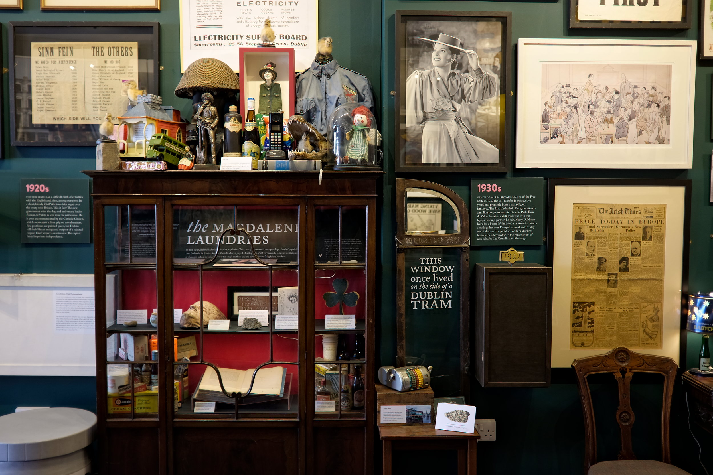A detailed museum display featuring a wooden cabinet labeled “The Magdalen Laundries” filled with historical artifacts, surrounded by vintage posters, photographs, clothing, and a tram window labeled “This window once lived on the side of a Dublin tram.”
