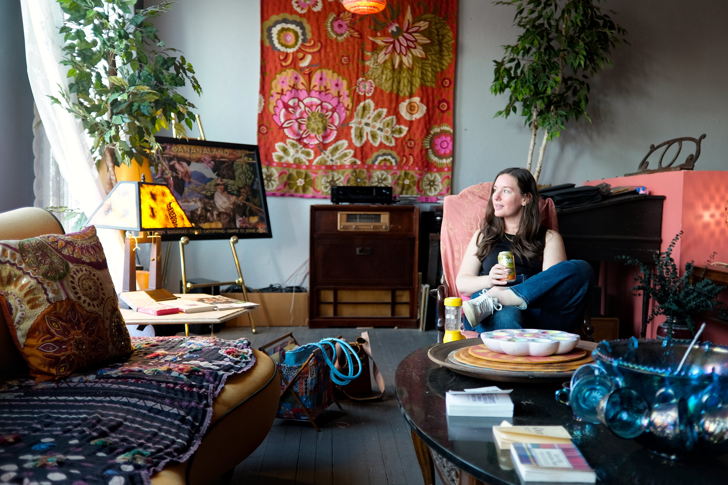 A woman with long brown hair sits in a pink armchair, holding a drink in a cozy, bohemian-style room decorated with vibrant textiles, potted plants, a vintage record player, and eclectic artwork including a framed “Bananaland” poster. The space features warm tones, patterned pillows, and a mix of modern and retro furniture, creating a relaxed, artistic vibe.