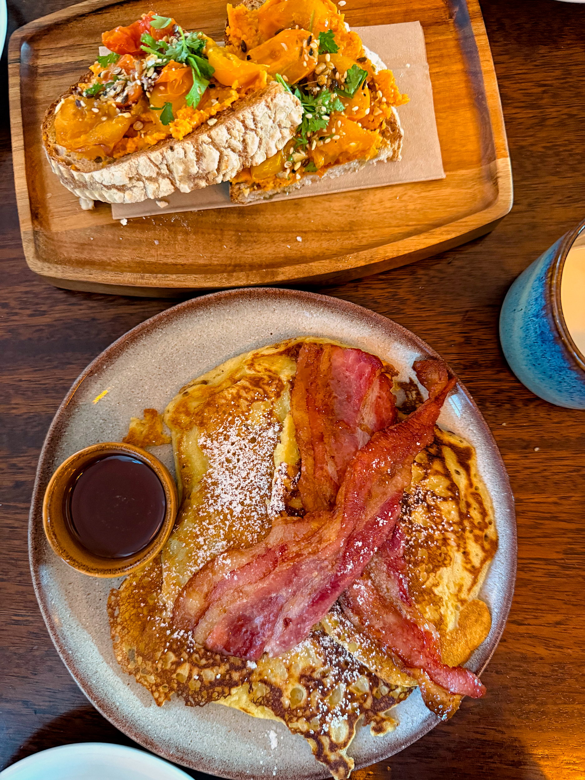 A plate with bacon and pancakes, and another with a hummus and tomato toast set on a wooden table at The Stop