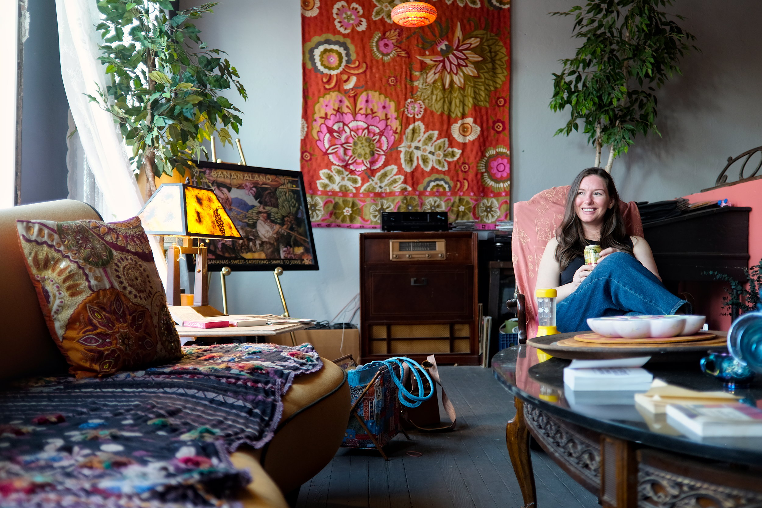 A woman with long brown hair sits smiling in a pink armchair, holding a drink in a cozy, bohemian-style room decorated with vibrant textiles, potted plants, a vintage record player, and eclectic artwork including a framed “Bananaland” poster. The space features warm tones, patterned pillows, and a mix of modern and retro furniture, creating a relaxed, artistic vibe.