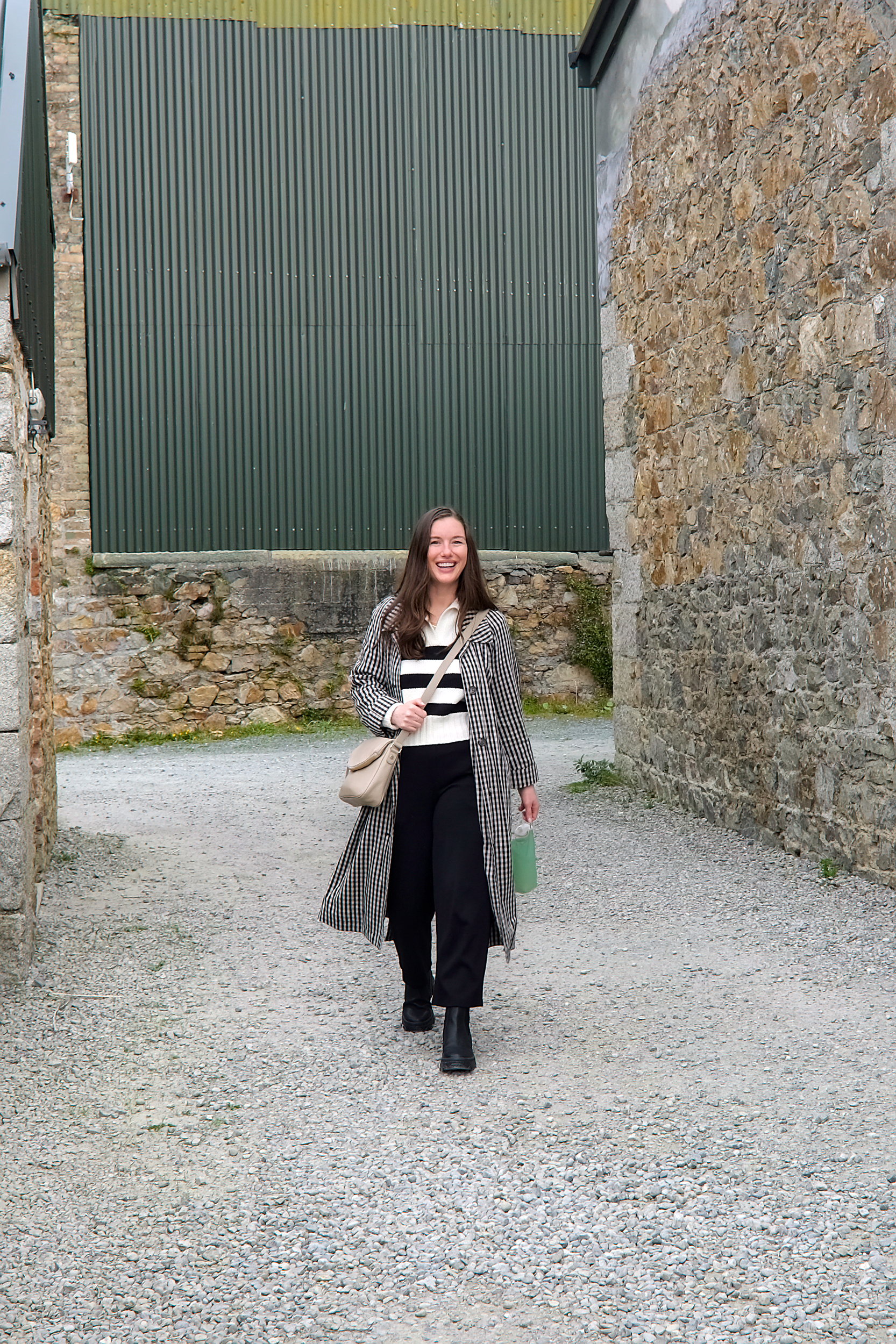 Alyssa walks through Kilruddery Yard wearing a striped polo, black pants, gingham trench, and black boots while carrying her water bottle