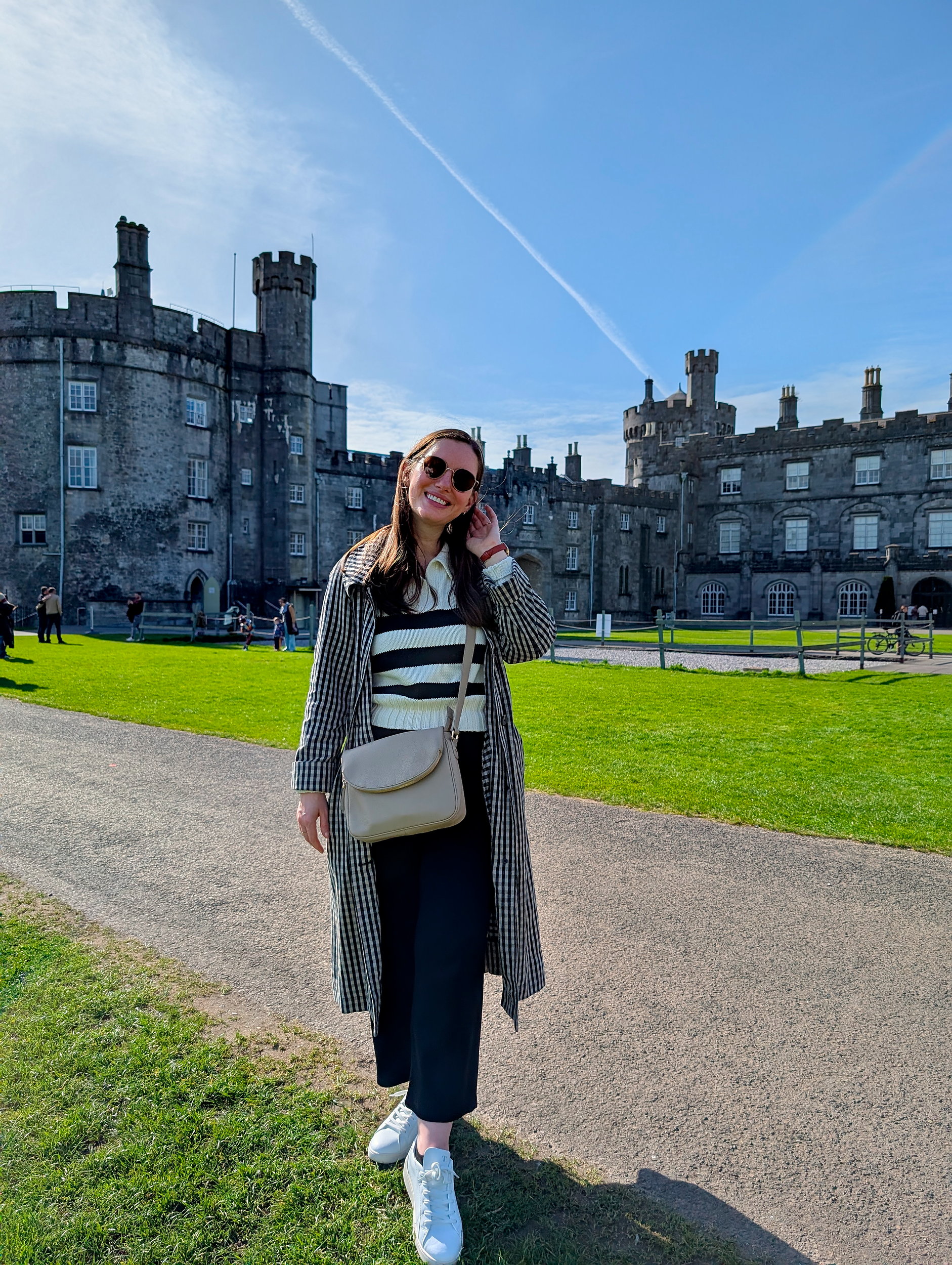 Alyssa in front of Kilkenny Castle, wearing a striped polo, gingham trench, culottes, and sneakers