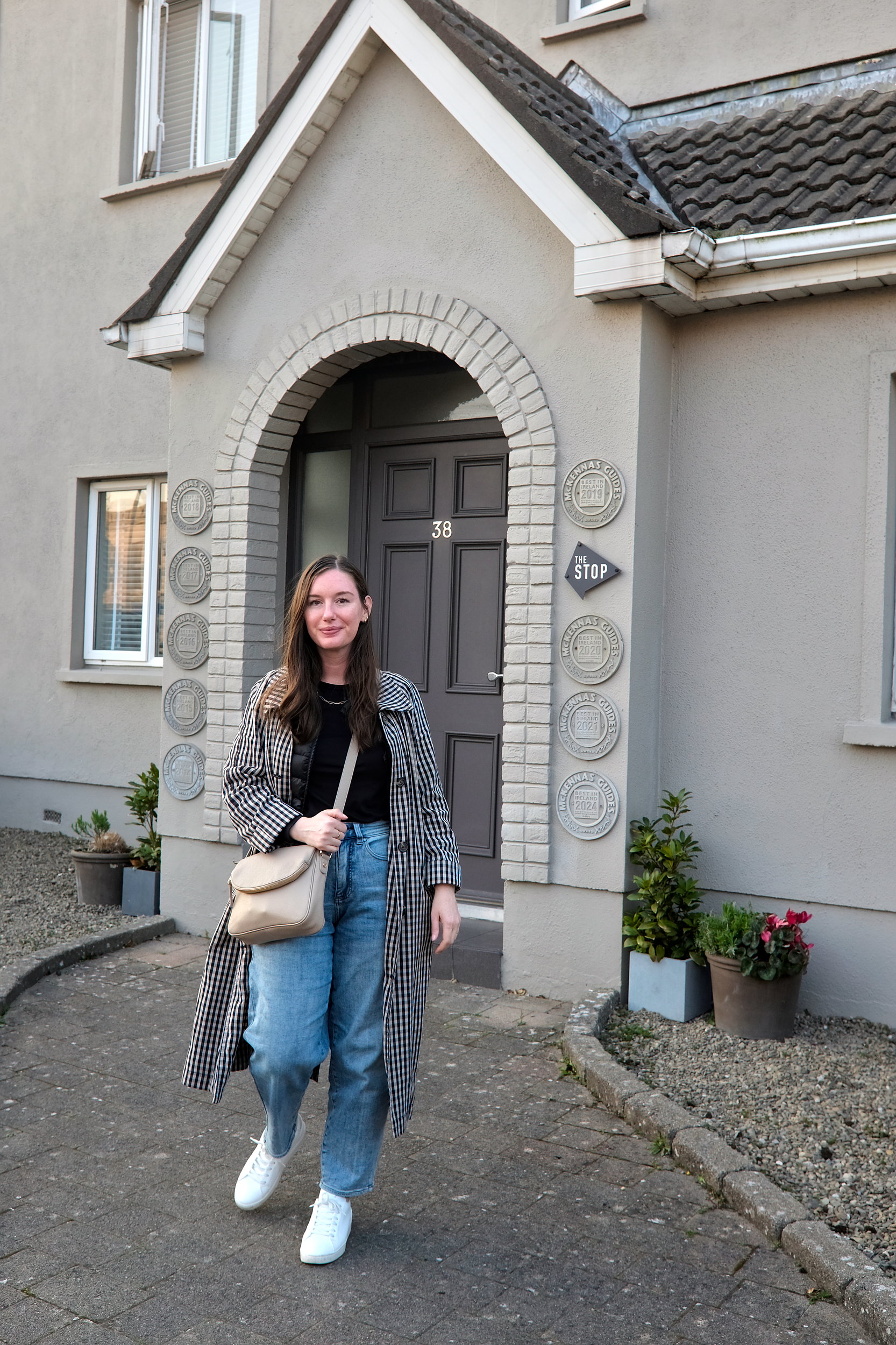 The author stands outside the entrance of The Stop Galway, a boutique bed and breakfast with a grey facade, arched brick doorway, and the number 38 above the door. Decorative award plaques and potted plants frame the stylish entryway.