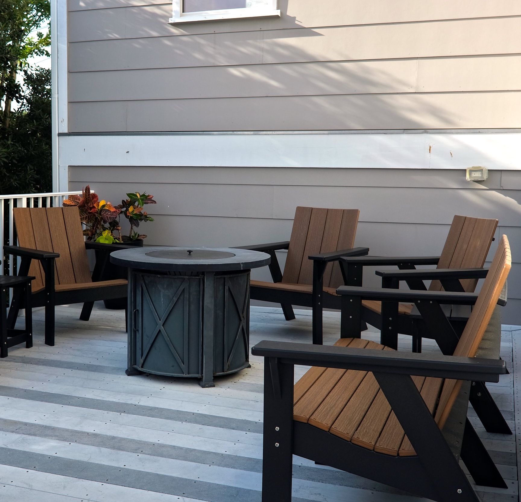 Cozy outdoor seating area with four wooden and black metal chairs arranged around a round metal fire pit on a striped wooden deck, set against a beige-paneled wall with a planter of colorful foliage nearby.